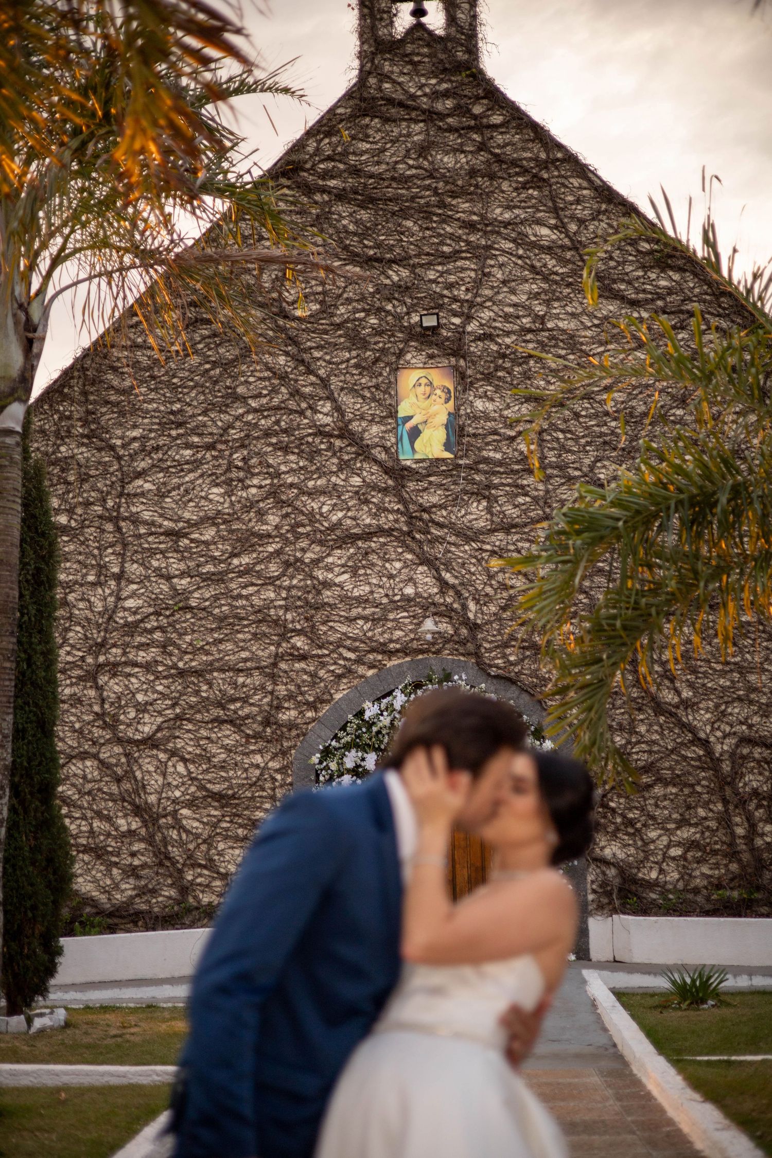 Bride and groom share a romantic kiss in front of a vine-covered chapel with a religious mosaic.
