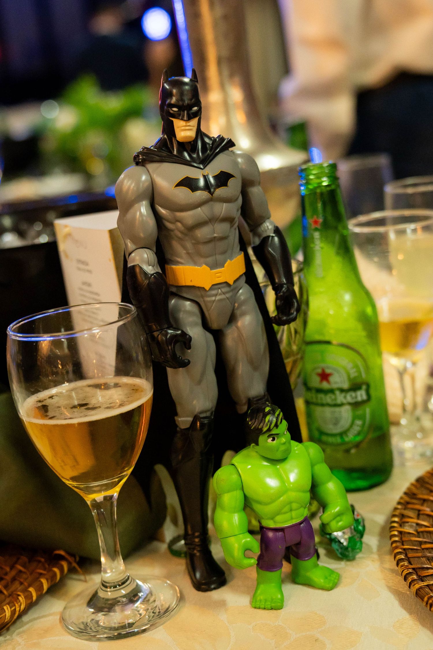 Batman and Hulk action figures posed on a table beside a Heineken beer bottle and a glass of beer.