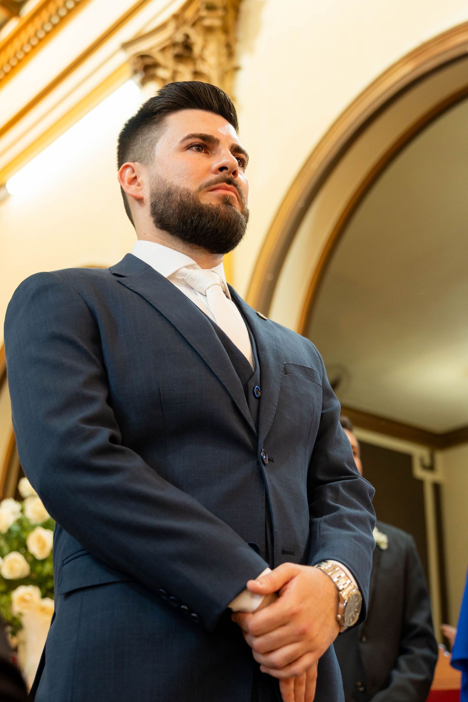Elegant bearded man in navy blue three-piece suit with watch, standing in ornate golden interior.