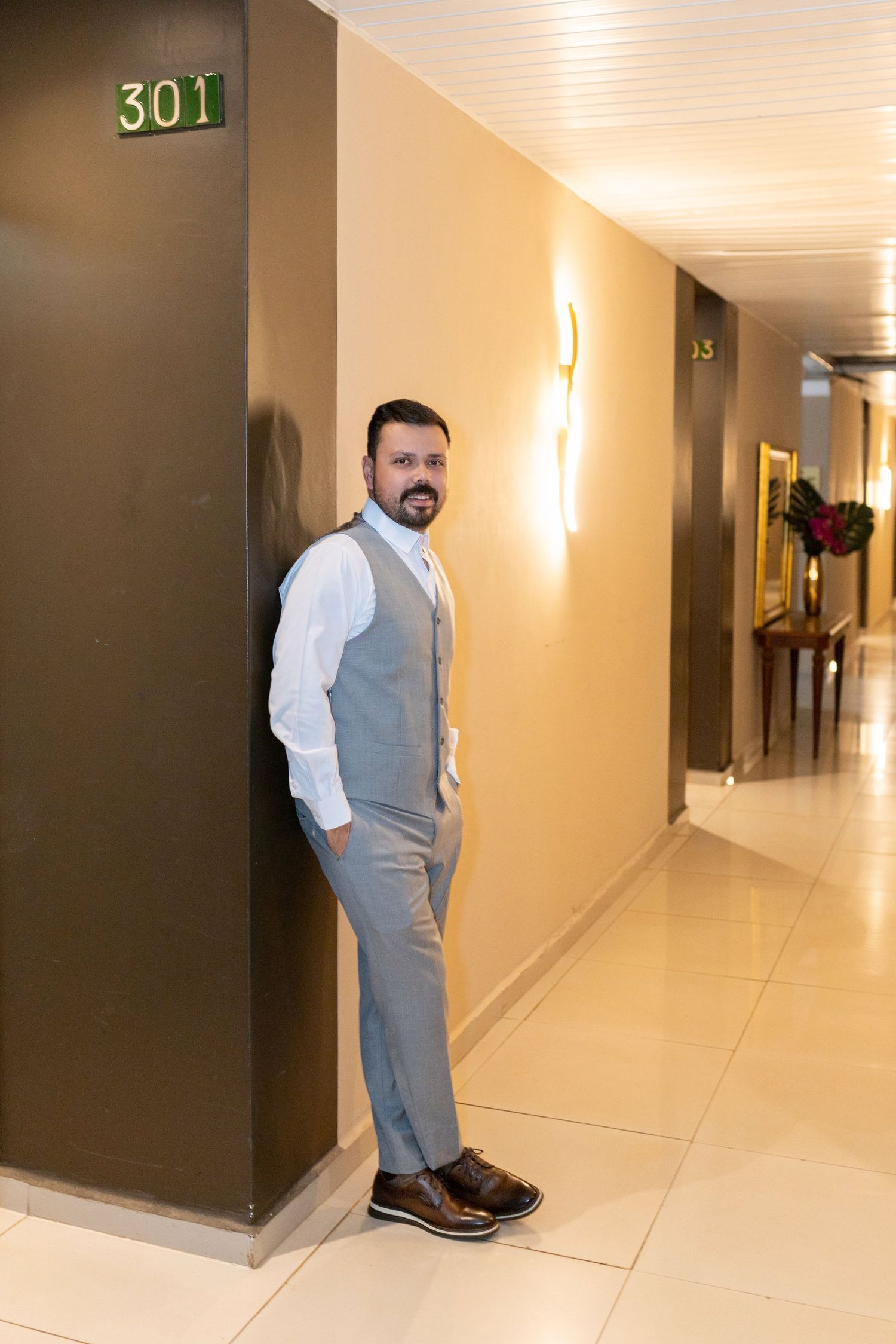 Smiling bearded man in gray vest and white shirt leaning against hotel corridor wall near room 301.