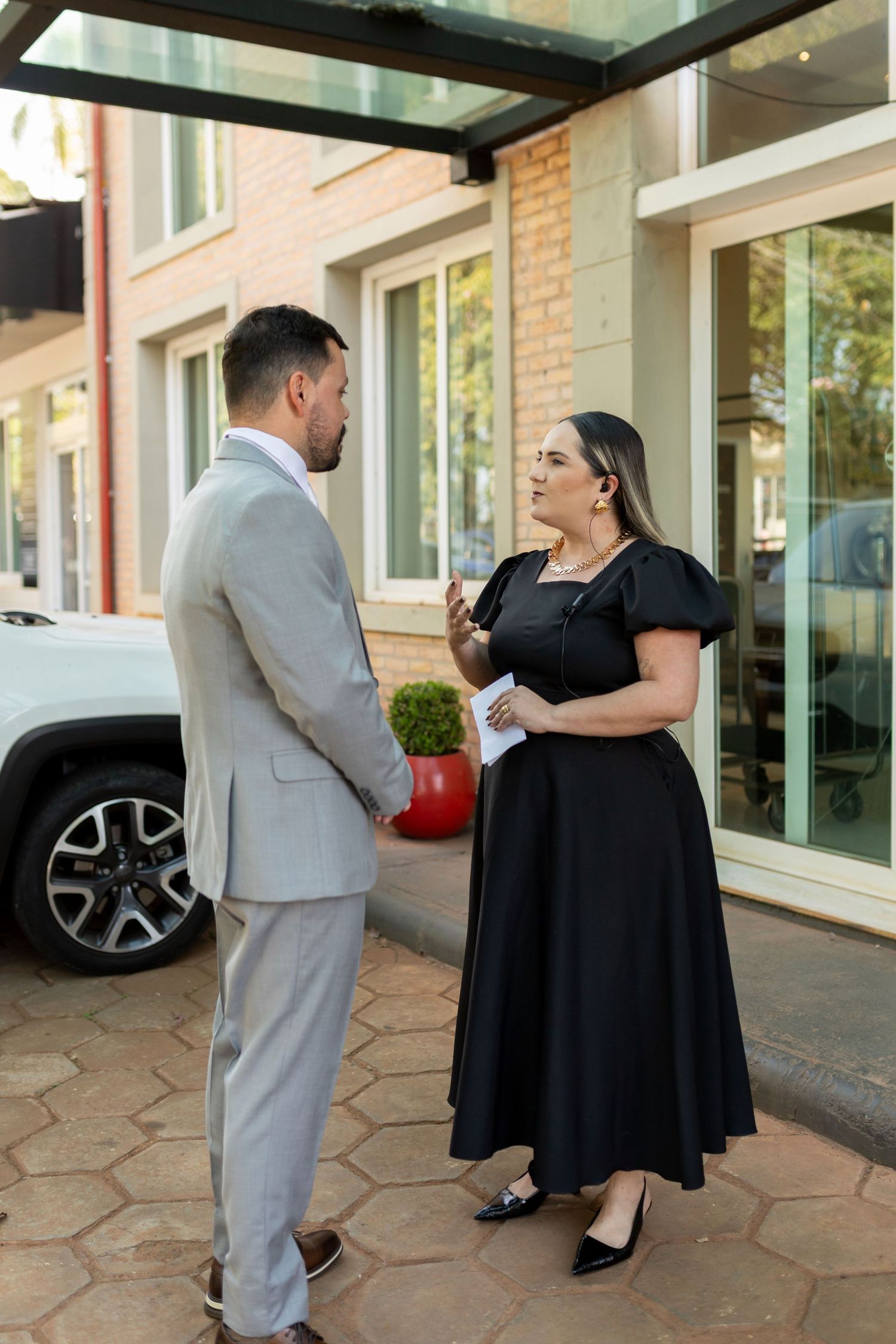 A man in a gray suit and a woman in a black midi dress have a conversation outside a modern building.