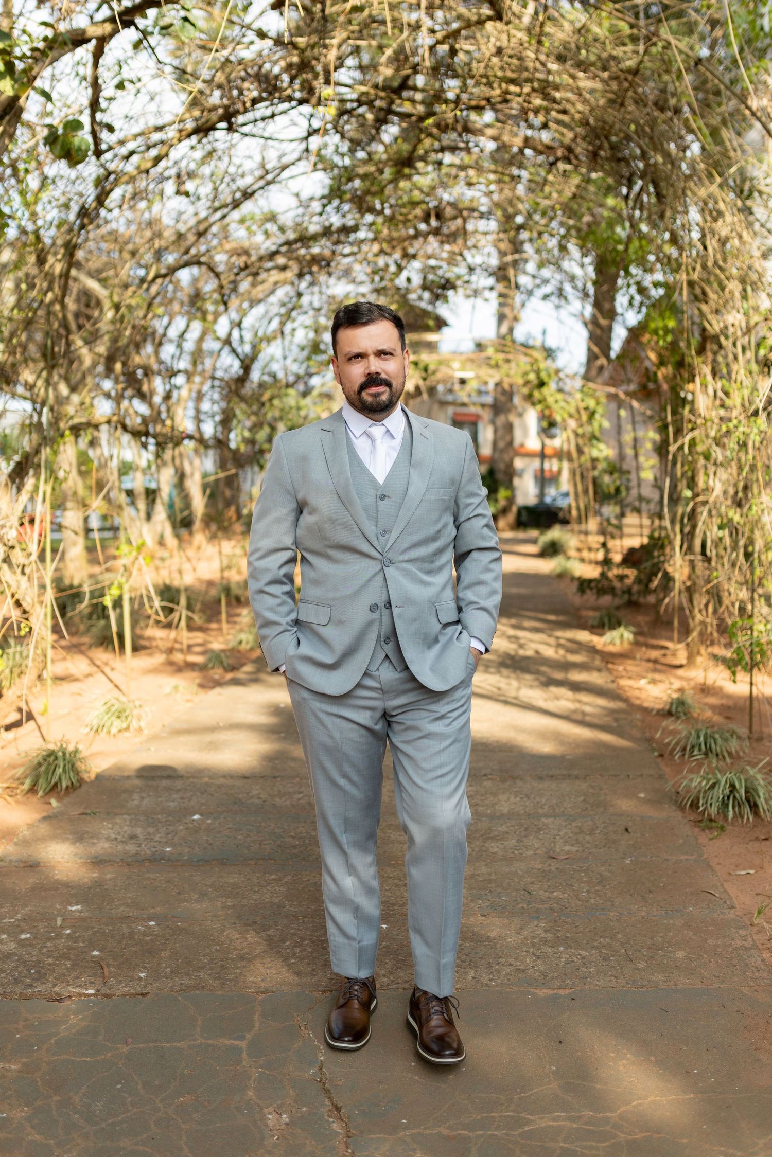 Bearded man in light gray three-piece suit with brown shoes standing on garden path under leafy archway.