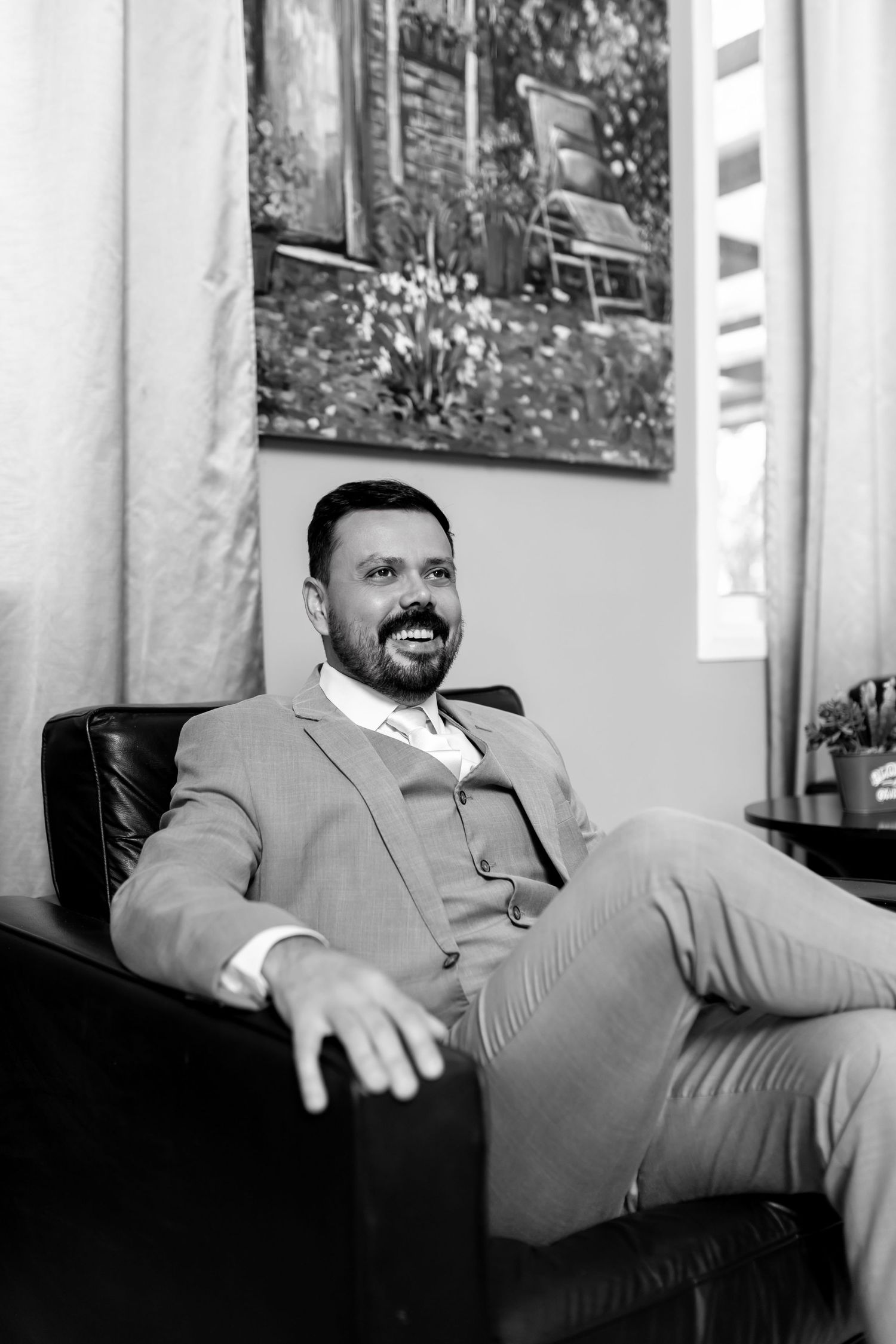 Smiling bearded man in a tailored suit relaxing in a leather chair, black and white portrait photo.