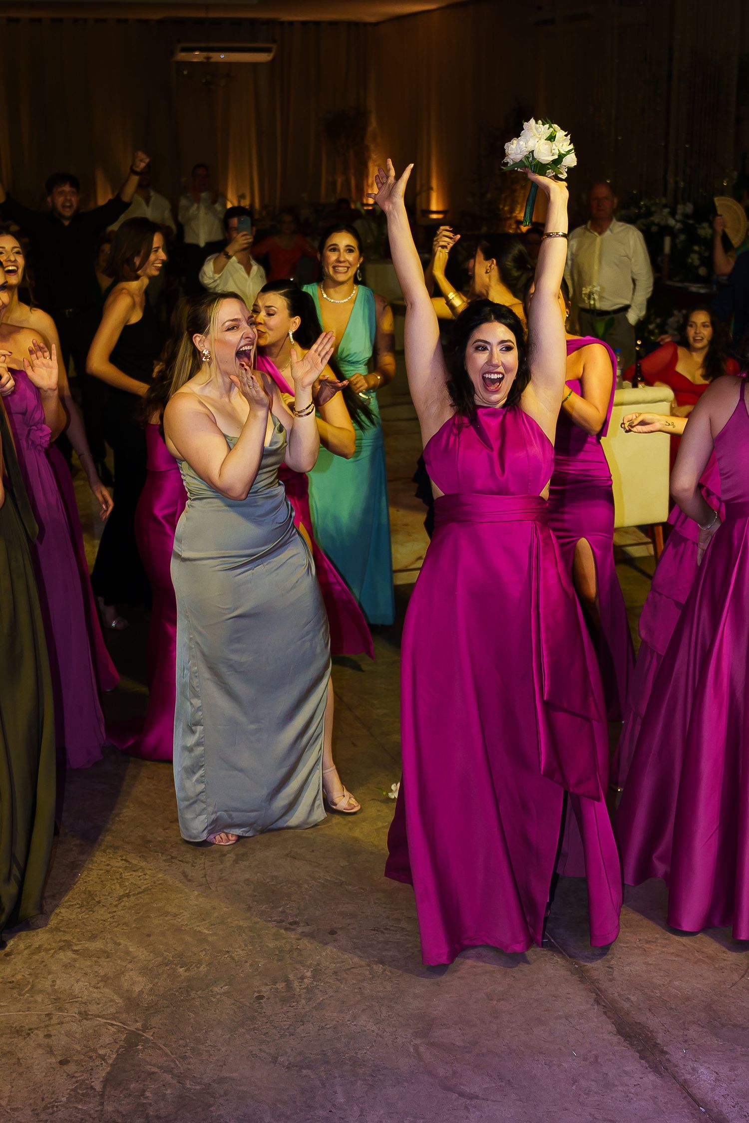 Women in magenta gowns celebrate at a wedding reception, one triumphantly holding a bouquet above her head.