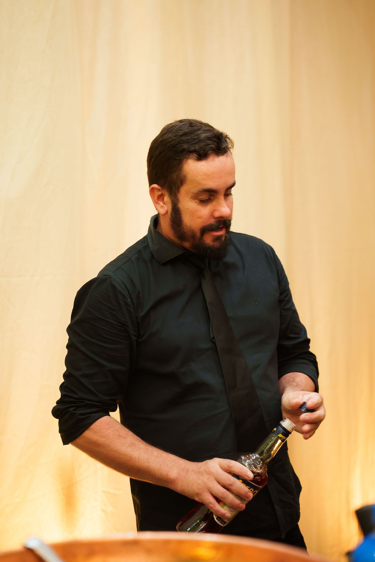 Bearded man in black shirt and tie opening a bottle at an event.
