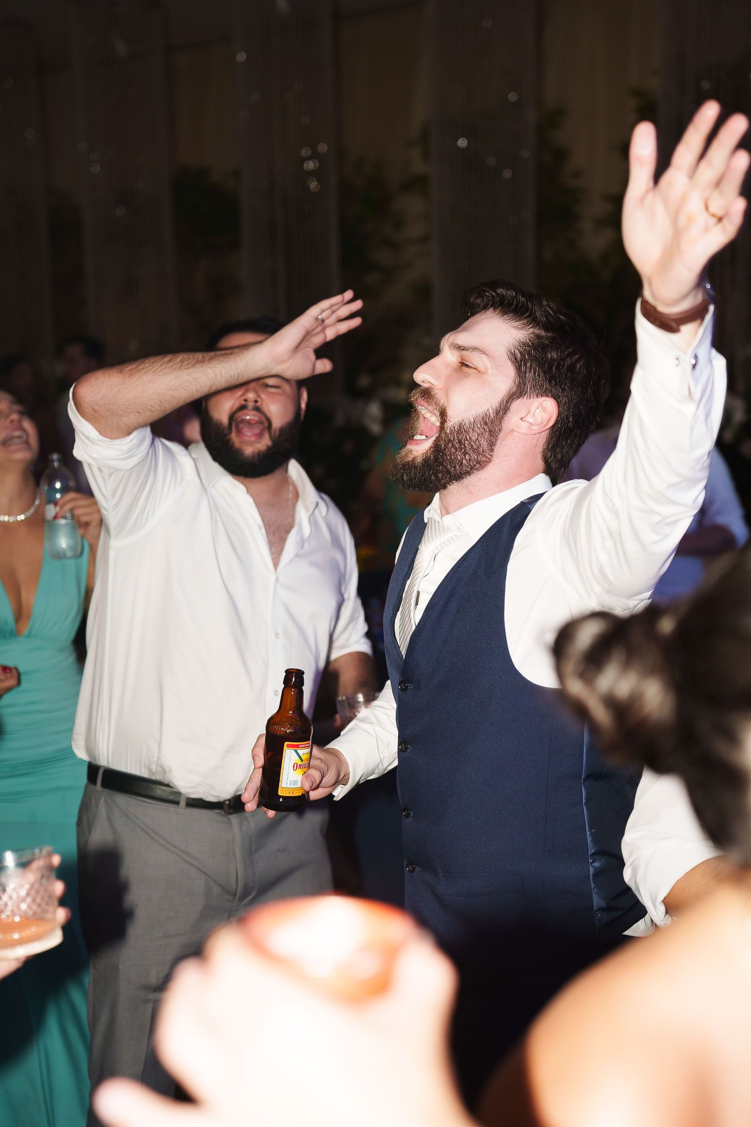 Two bearded men dancing joyfully at a wedding reception, one holding a beer bottle, surrounded by guests.