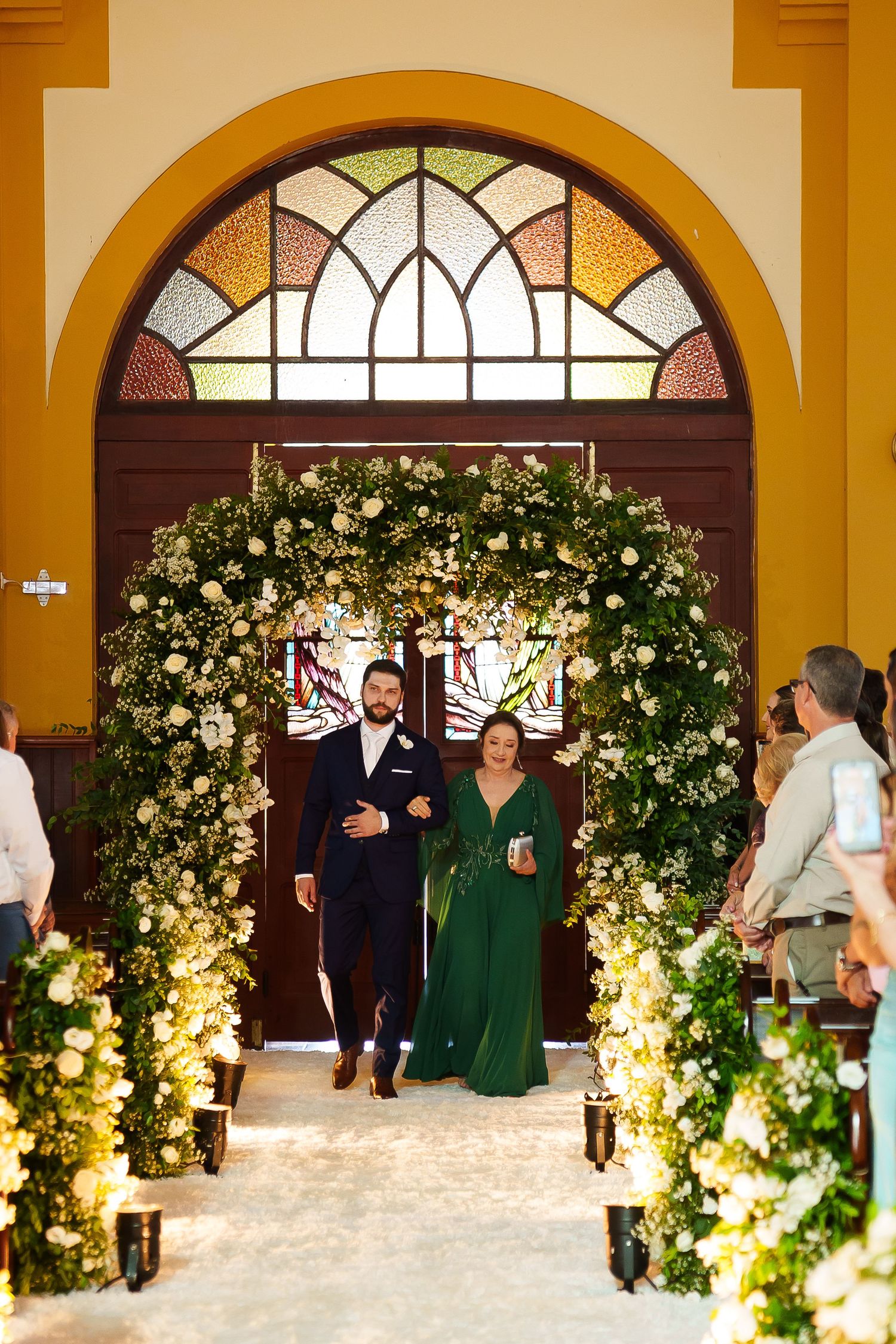 Couple walks down floral arch aisle at elegant church wedding ceremony with white flowers and greenery.