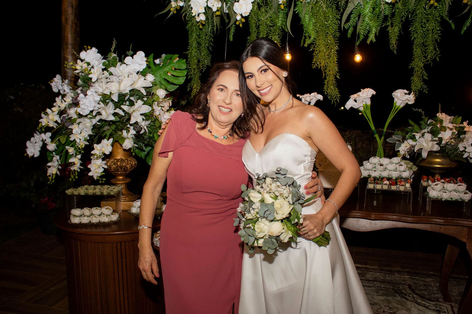 Bride in white gown holding bouquet poses with woman in pink dress at elegant floral wedding reception.