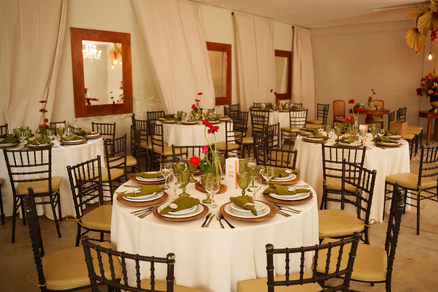 Elegant banquet hall with round tables set with white linens, gold chargers, and red floral centerpieces on chiavari chairs.