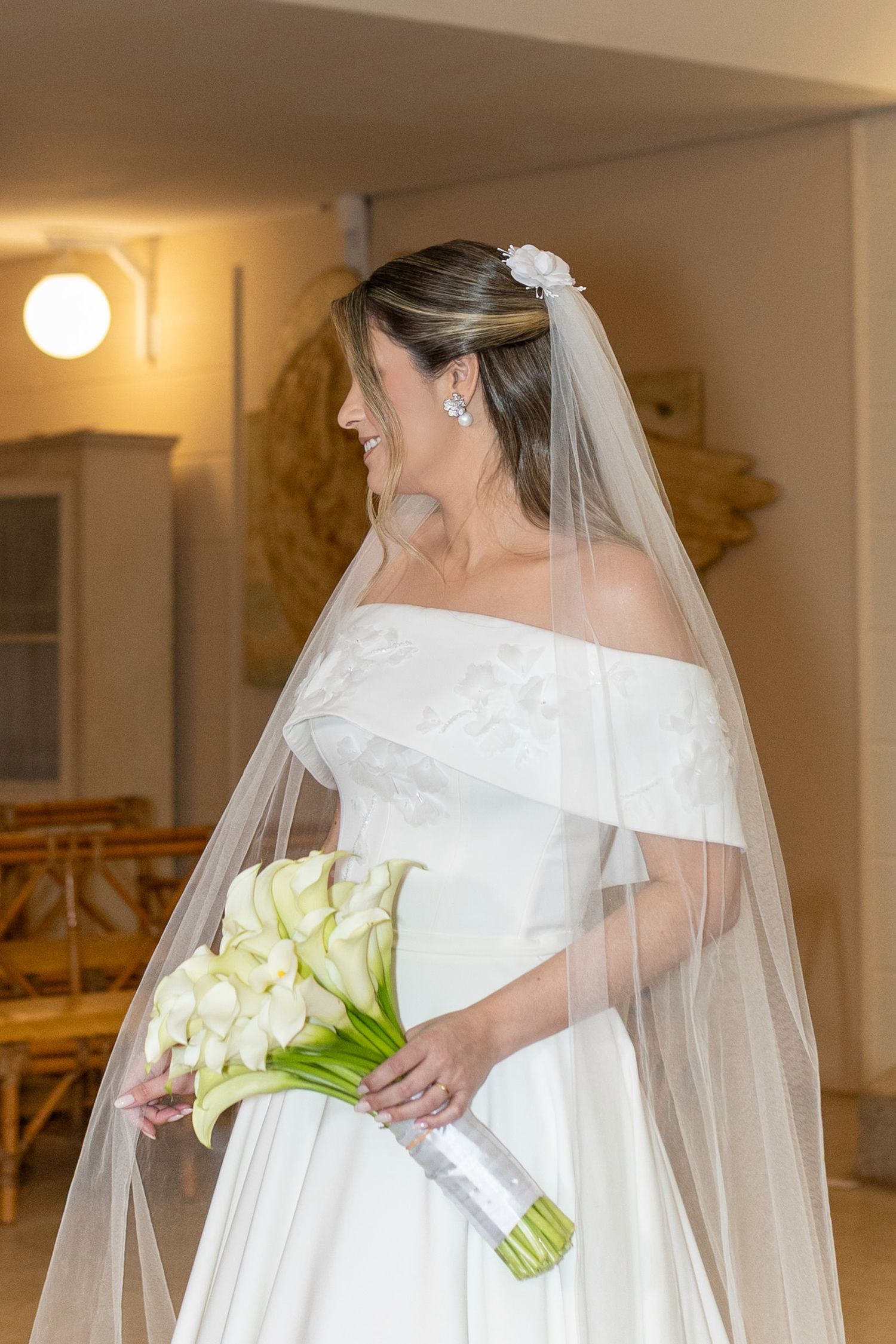 Smiling bride in off-shoulder white gown and veil holds calla lily bouquet indoors.