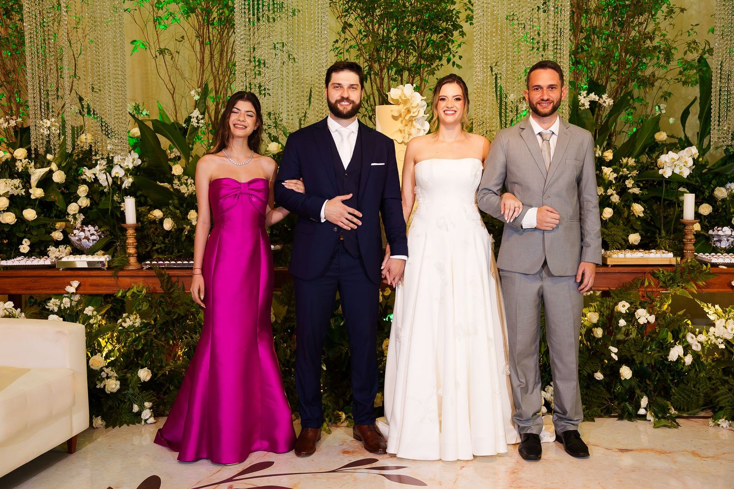 Wedding party of four posing before floral backdrop, bride in white gown, groom in navy suit, guests in fuchsia dress and gray suit.