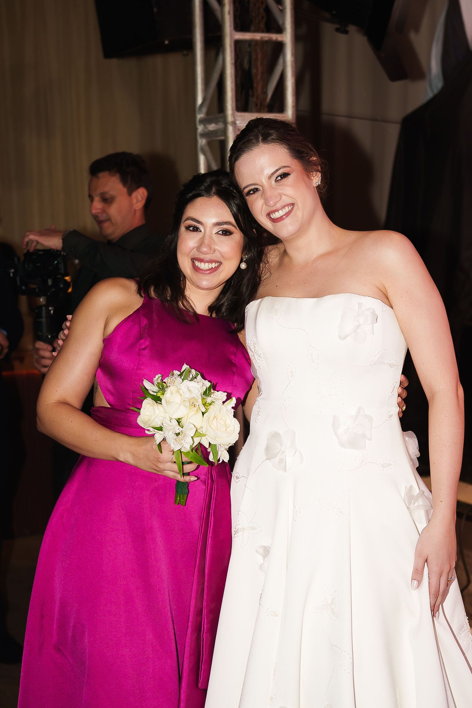 Bride in white strapless gown poses with bridesmaid in fuchsia dress holding white bouquet at wedding reception.