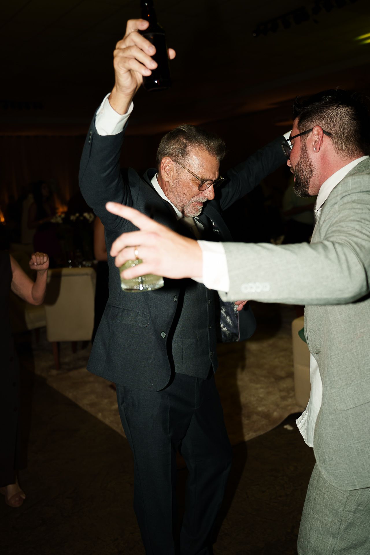 Two men in suits dancing joyfully at a wedding reception, one raising his hand with a drink.