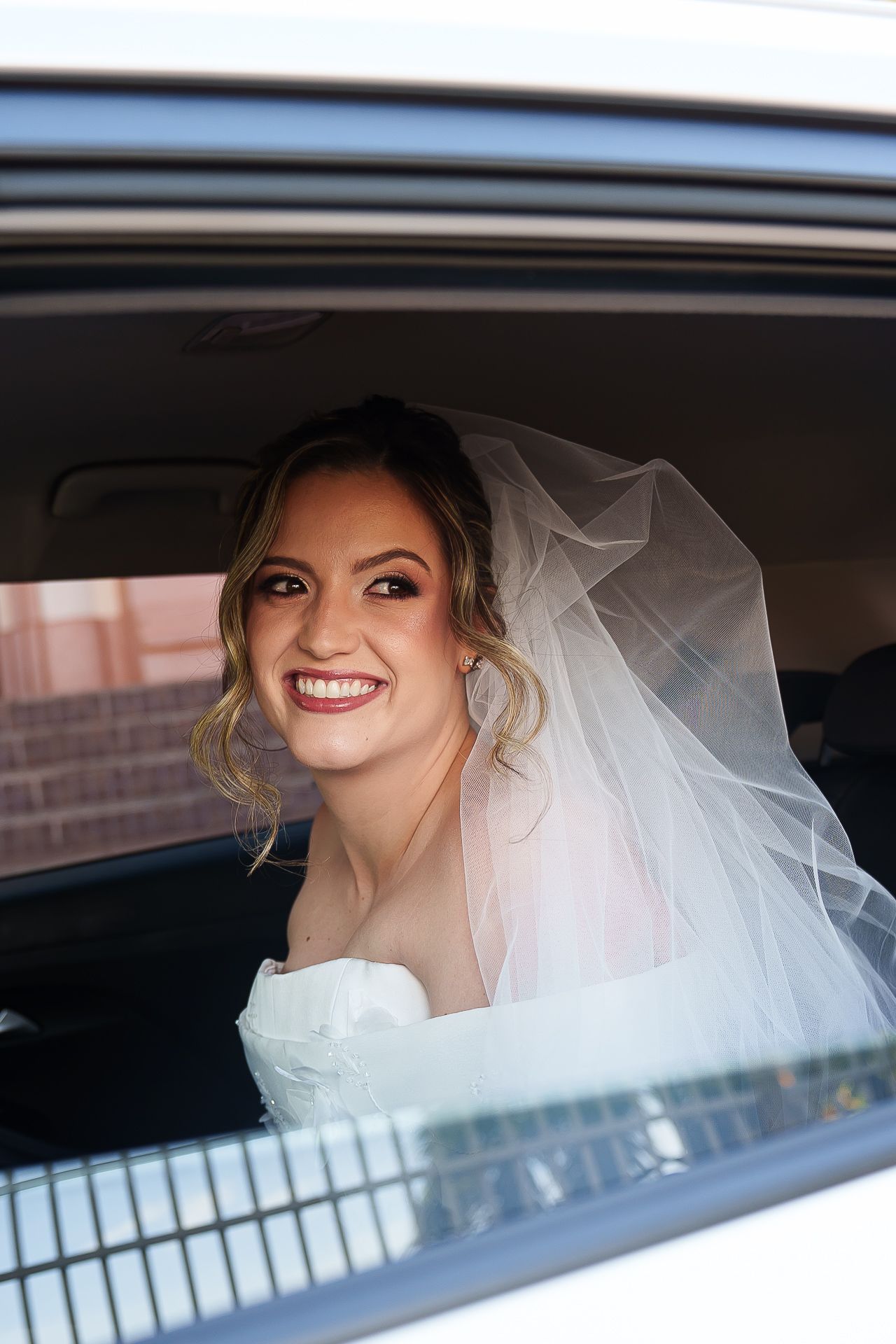 Smiling bride in white veil and gown looking out car window on wedding day.
