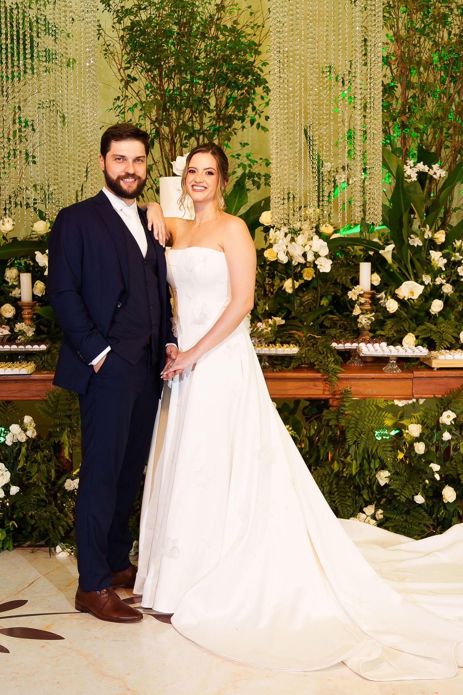 Bride in white strapless gown and groom in navy suit pose together at elegant wedding reception with floral backdrop.