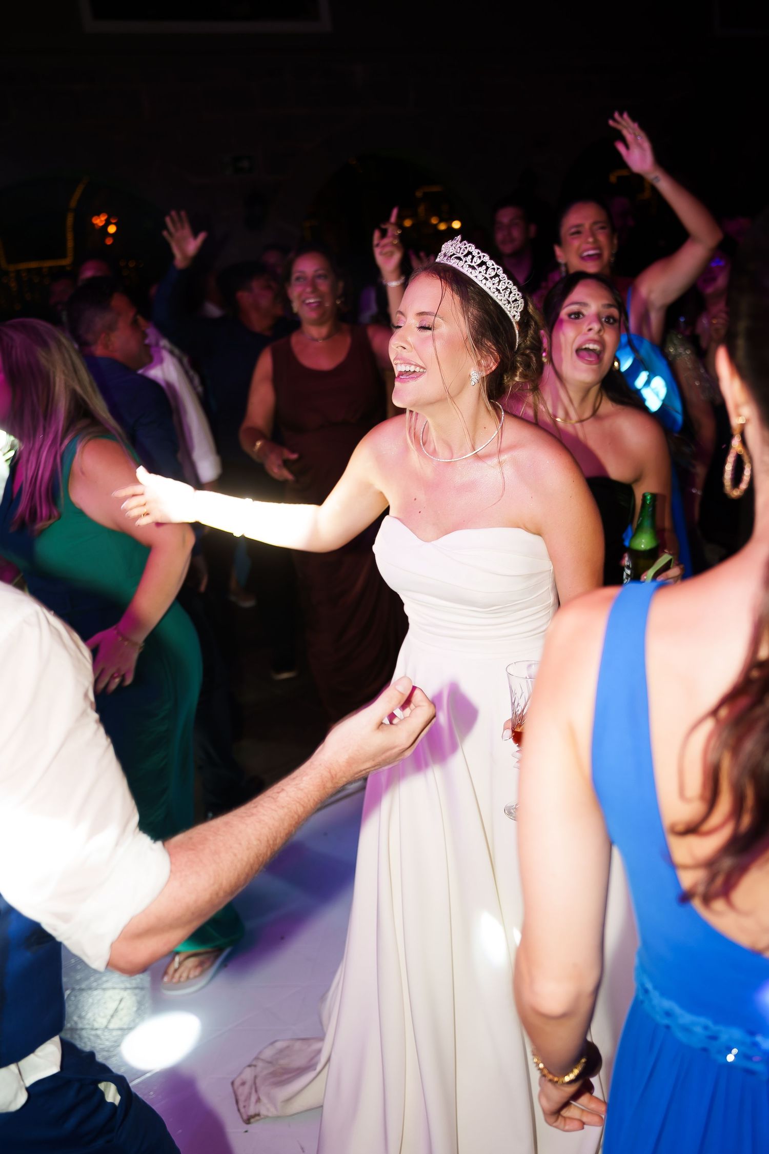 Bride in white gown and tiara dancing joyfully with guests at a lively wedding reception.