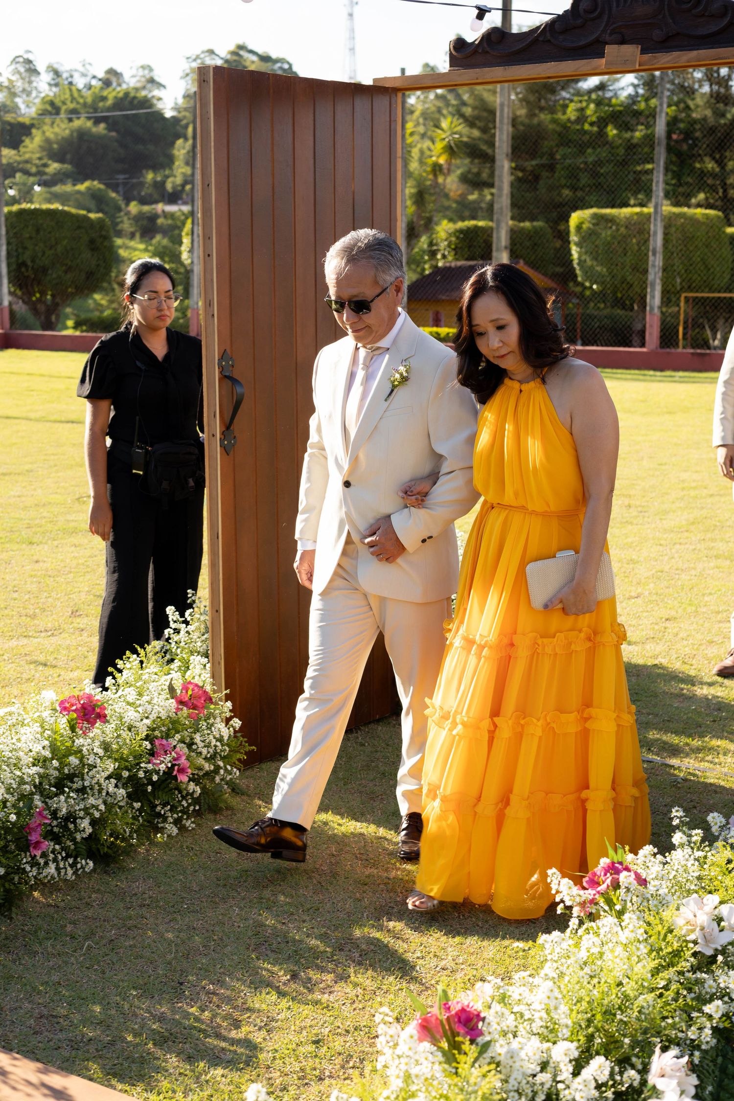 Elegant couple walking down floral aisle at outdoor wedding, man in white suit, woman in yellow tiered gown.