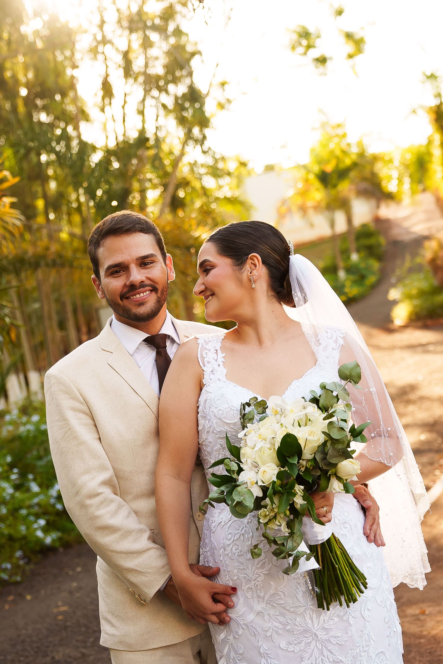 Smiling bride and groom pose outdoors in golden sunlight, she holds white bouquet, wearing lace gown and veil, he in tan suit.