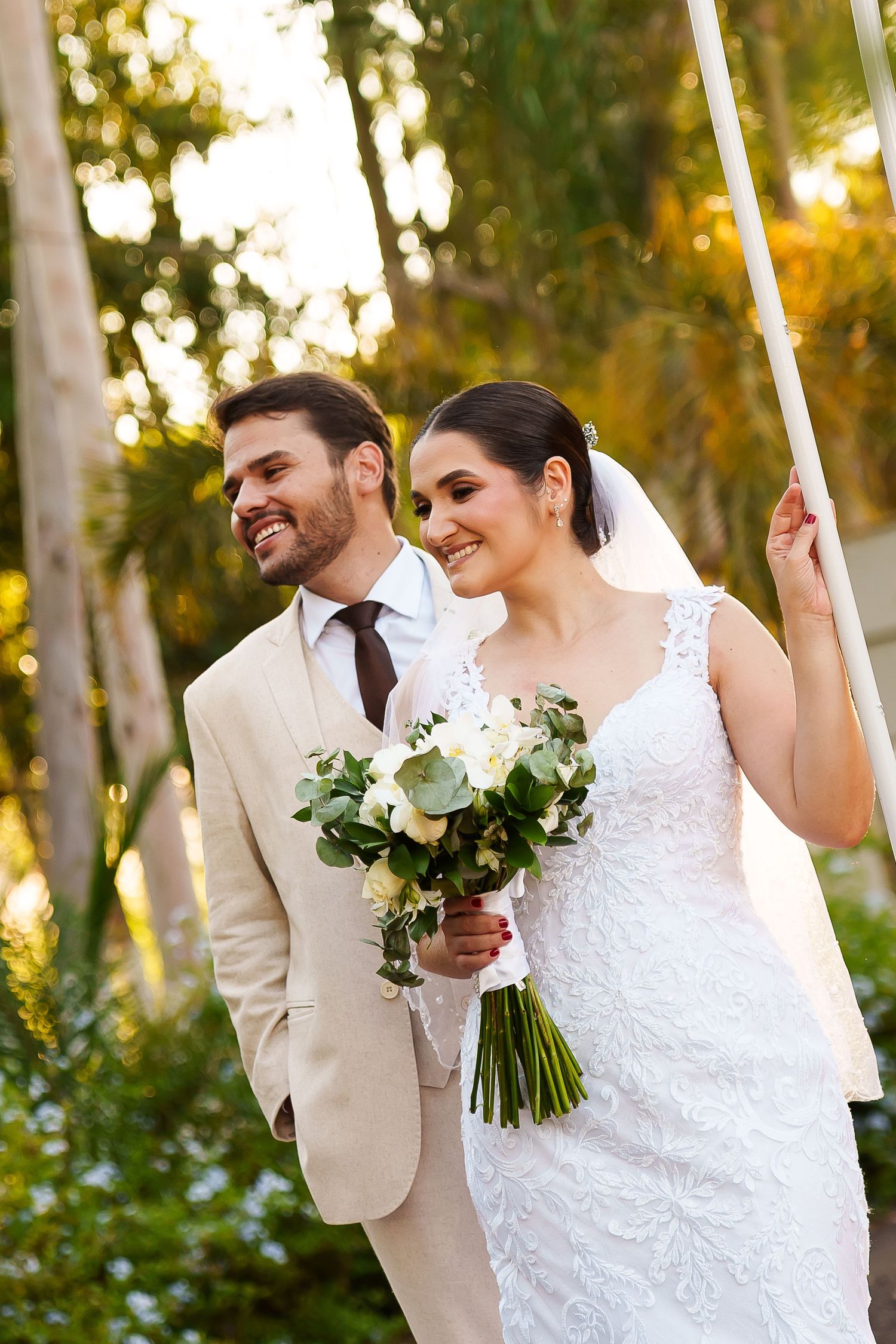 Smiling bride in lace gown holding white bouquet stands with groom in tan suit outdoors among green trees.