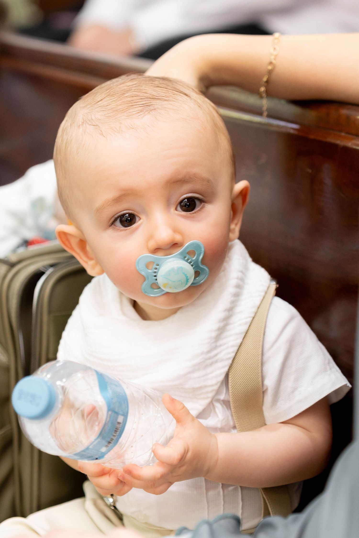 Cute baby with blue pacifier holding a water bottle, wearing a white bib and shirt, sitting near luggage.