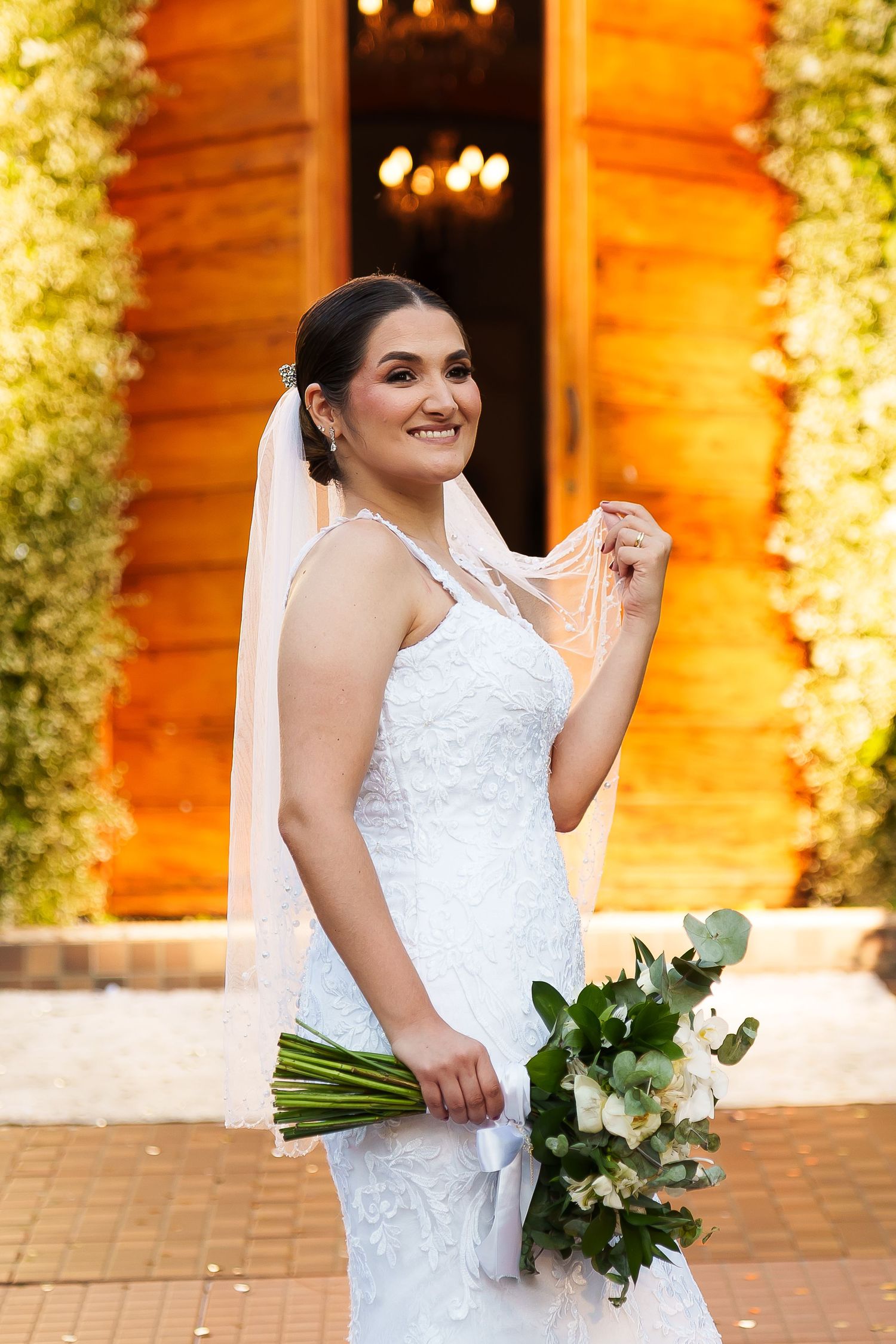 Smiling bride in white lace gown holding bouquet of white flowers, posing outdoors near wooden chapel.