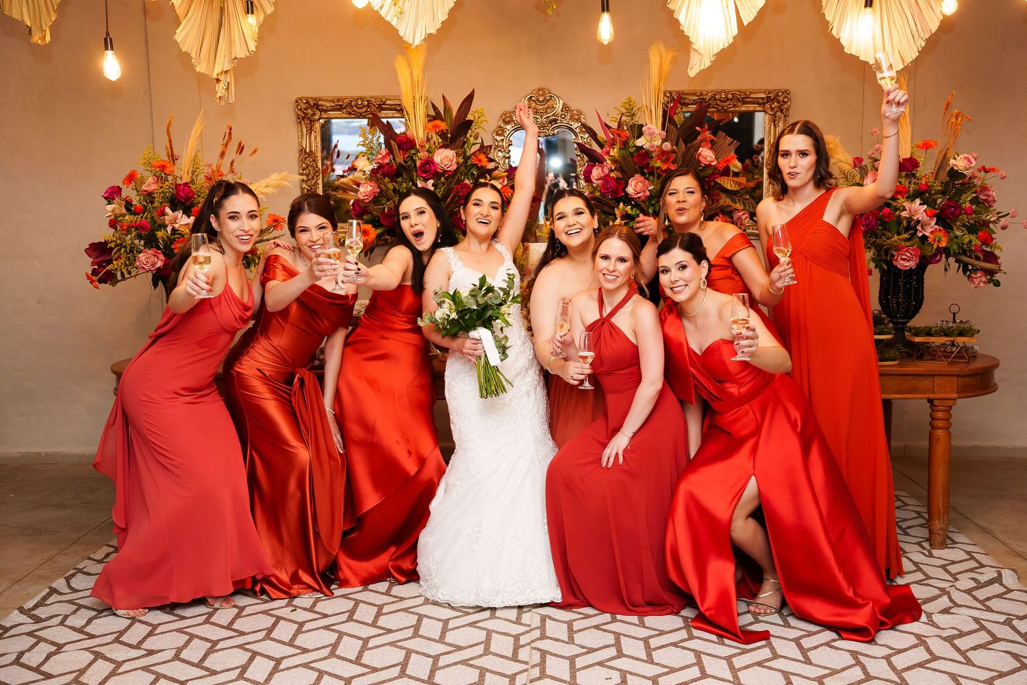 Bride in white gown poses with seven bridesmaids in red satin dresses, holding bouquets and champagne glasses indoors.