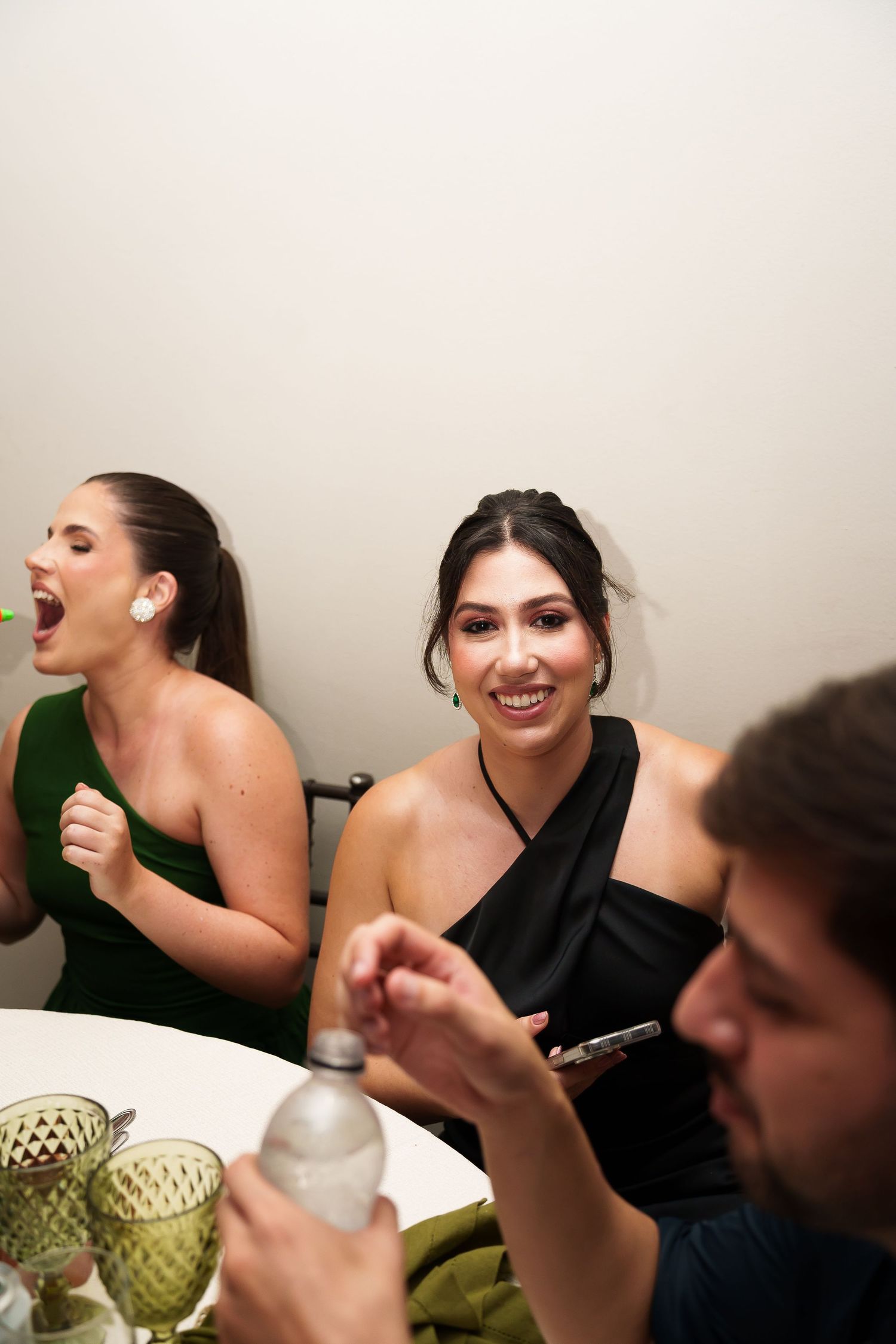 Two women in elegant dresses laughing at a dinner party, one in green, one in black, with glasses on the table.