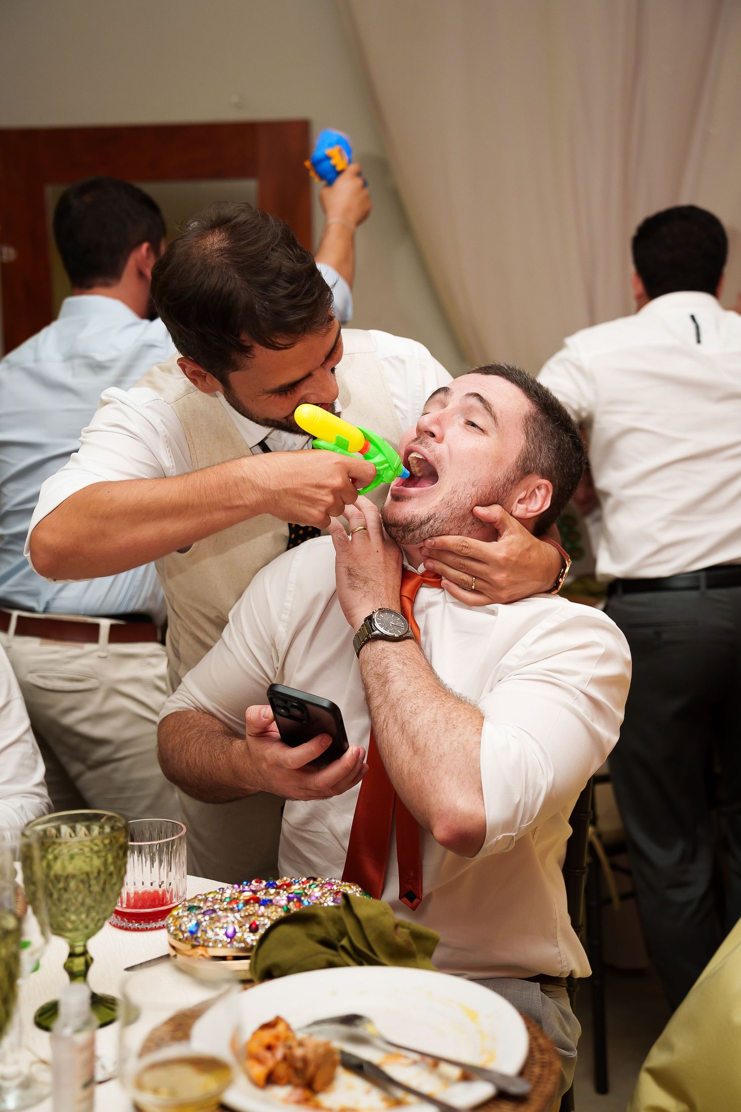 Men in white shirts celebrate at a party, one squirting a toy water gun into another's mouth at a festive dinner table.
