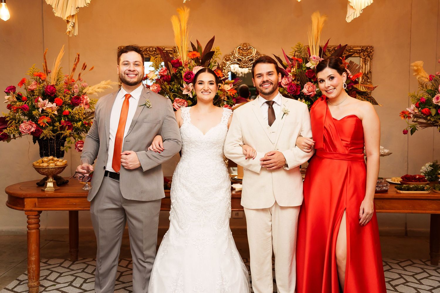 Two couples pose at a wedding, bride in white gown, groom in cream suit, guests in gray suit and red dress.