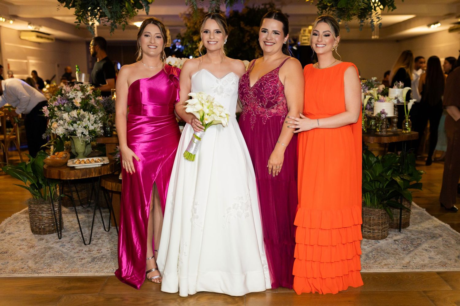 Bride in white gown poses with three women in colorful pink, magenta, and orange gowns at elegant wedding reception.