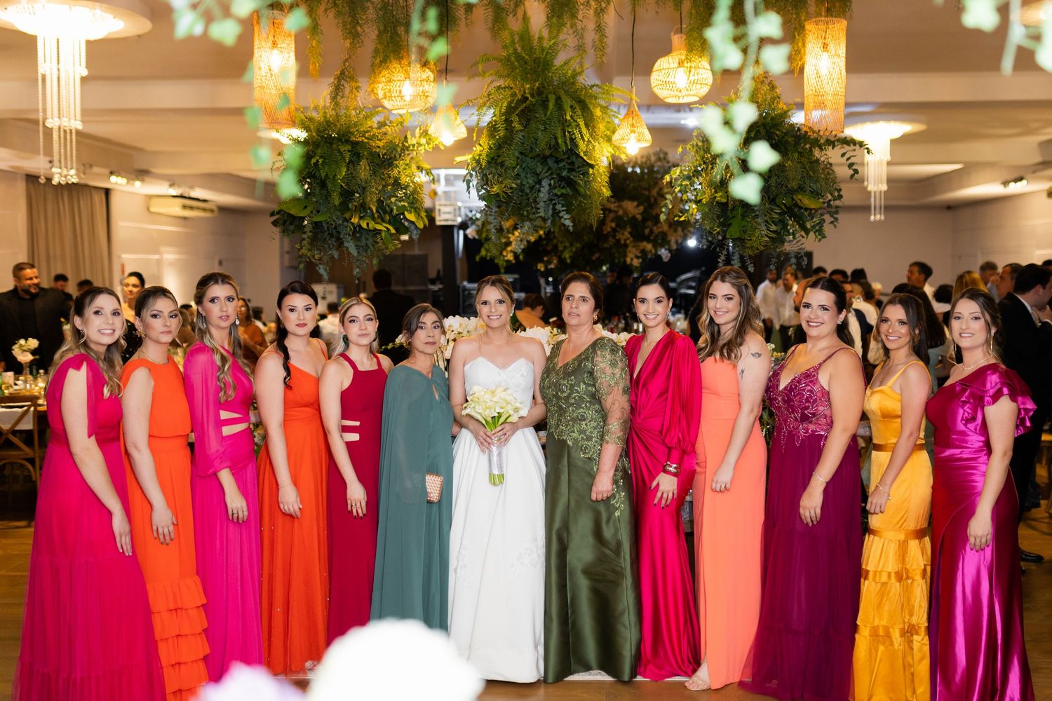 Bride poses with bridesmaids in colorful rainbow gowns at elegant wedding reception with lush greenery.