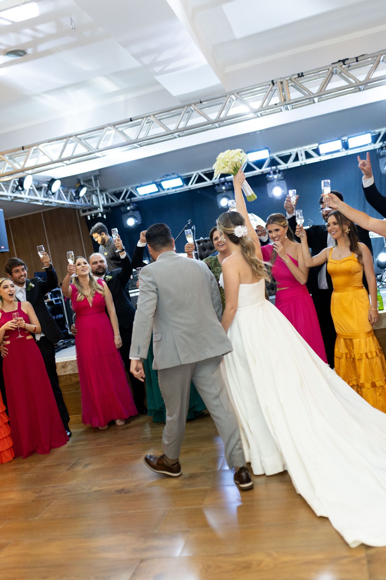 Bride and groom share first dance surrounded by guests in colorful gowns raising glasses at elegant wedding reception.