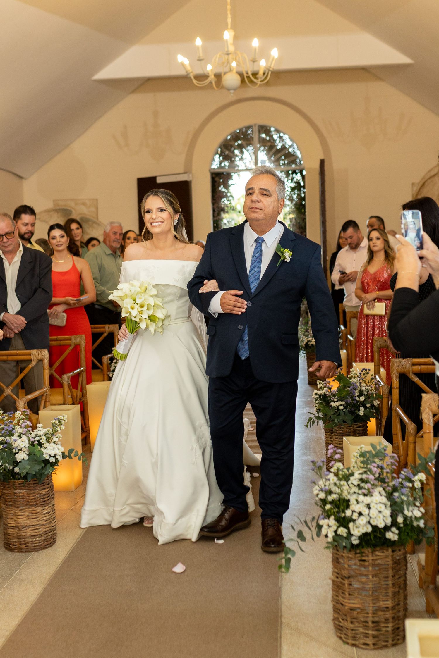 Bride in white gown walks down chapel aisle with escort, flanked by floral arrangements and seated guests.