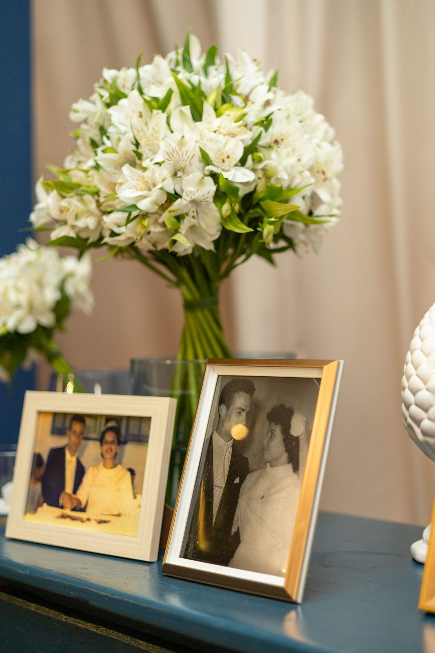 Wedding photos in gold frames displayed on blue table with white alstroemeria bouquet in glass vase.