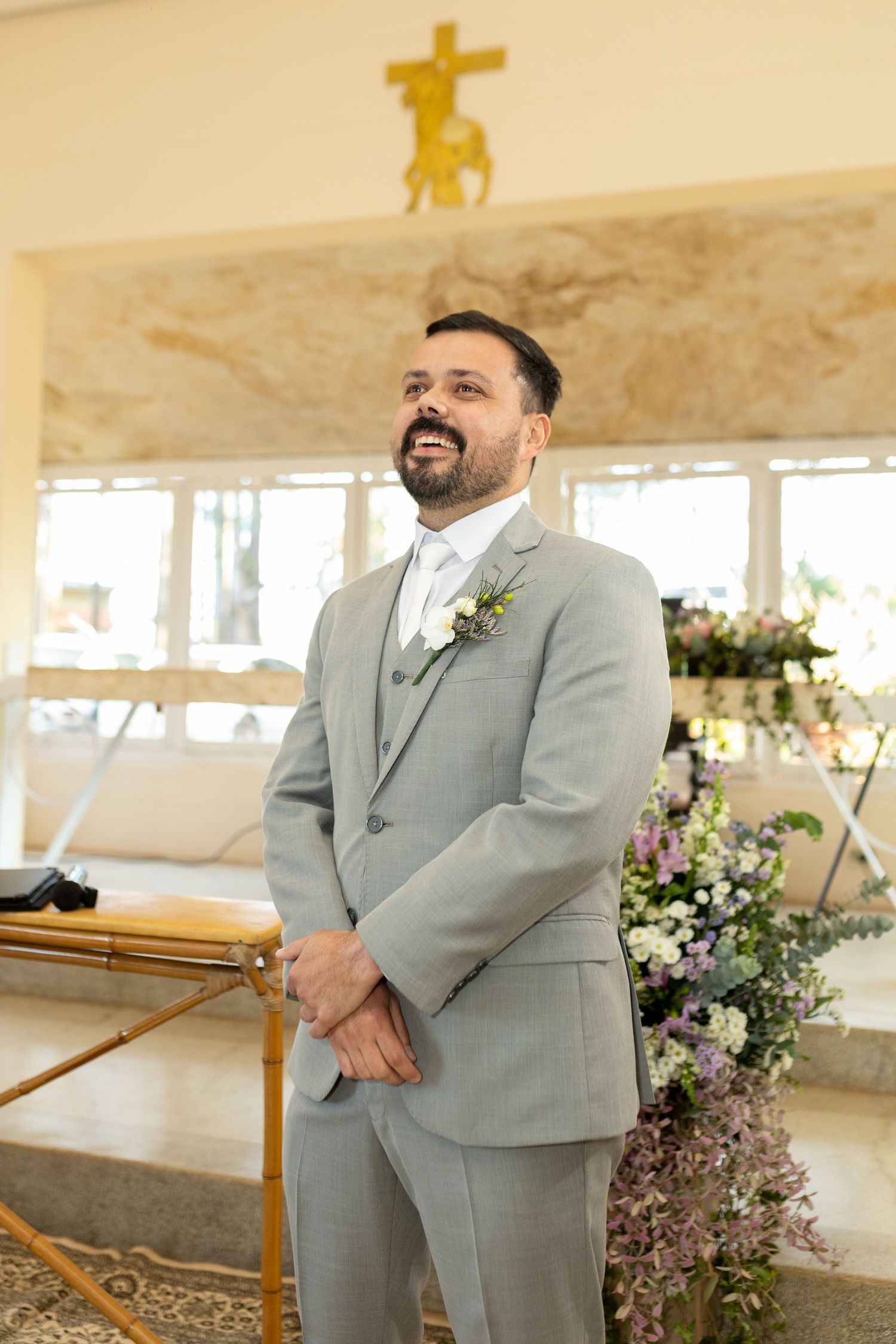 Groom in gray suit with boutonniere smiling at altar in decorated church during wedding ceremony.