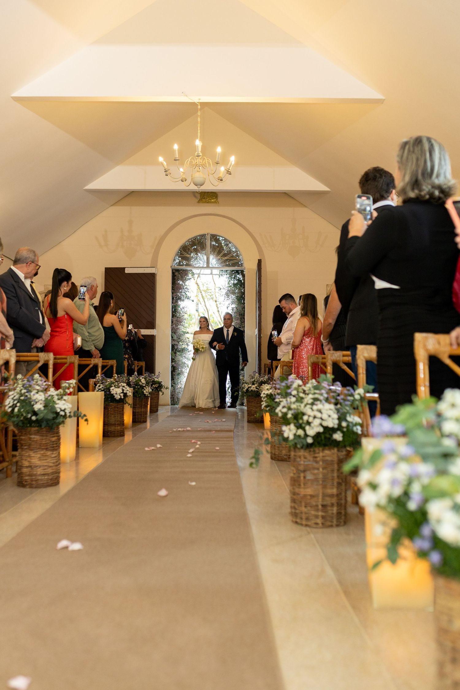 Bride walking down the aisle with escort in elegant chapel, guests capturing the moment, floral baskets lining the path.