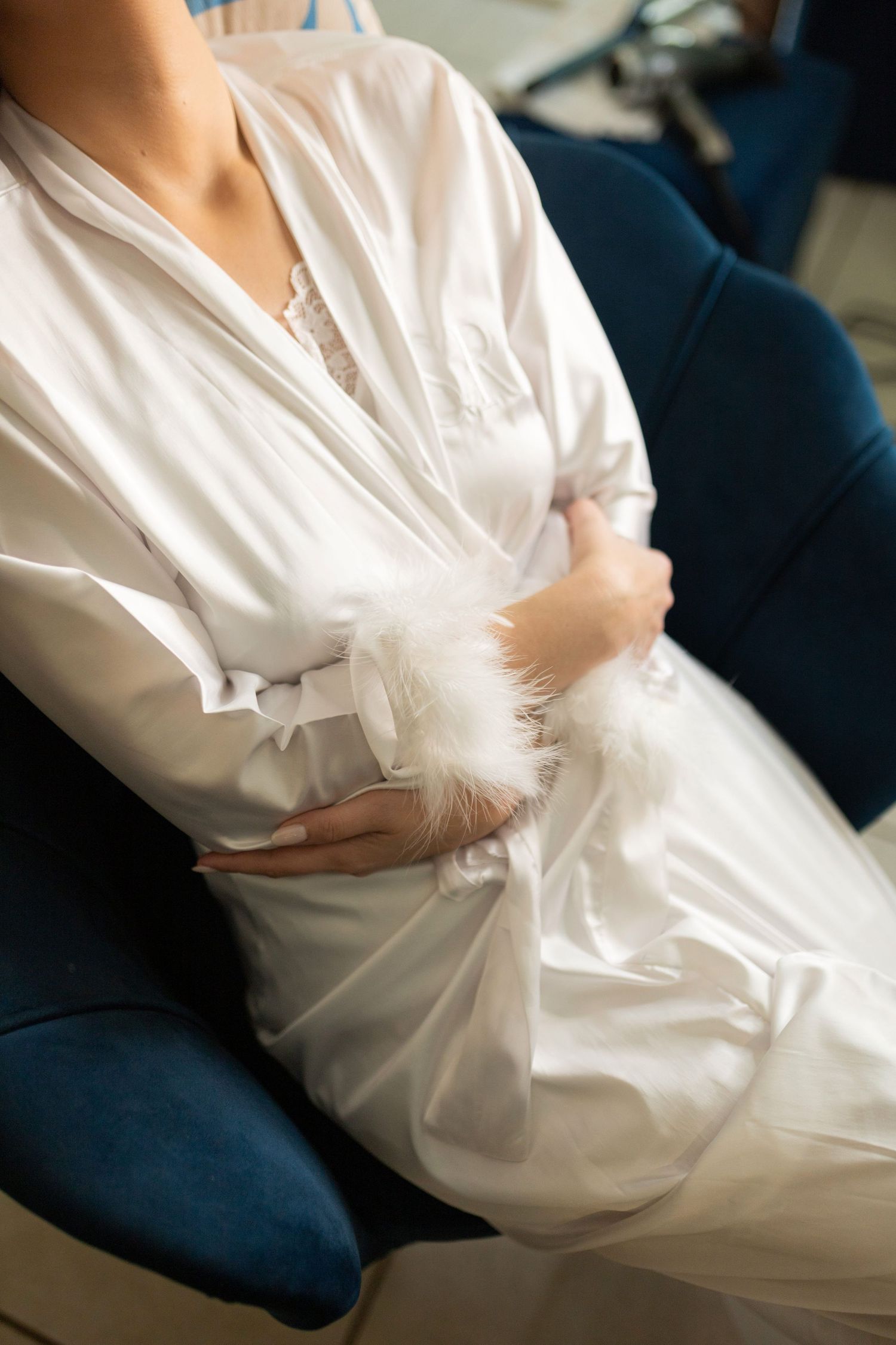 Bride in white satin robe holding feathered bouquet, sitting in blue velvet chair during wedding preparation.