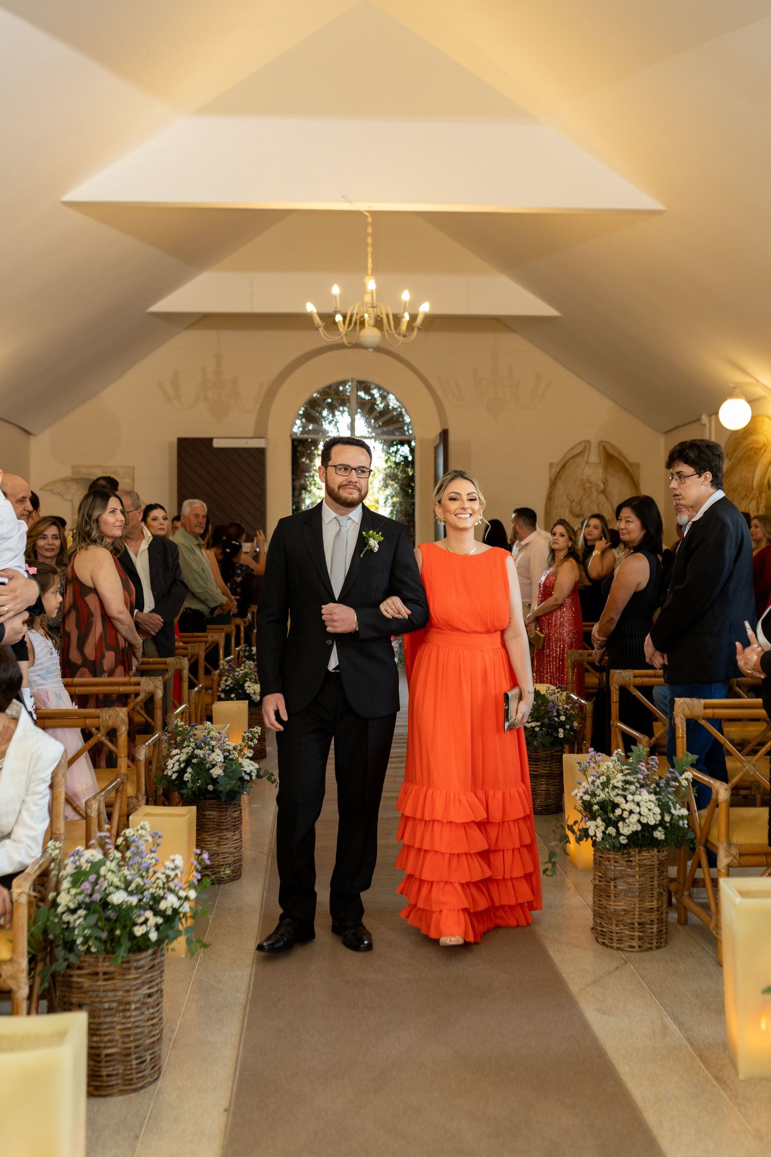 Couple walking down wedding aisle, woman in orange ruffled gown, man in dark suit, elegant chapel setting.