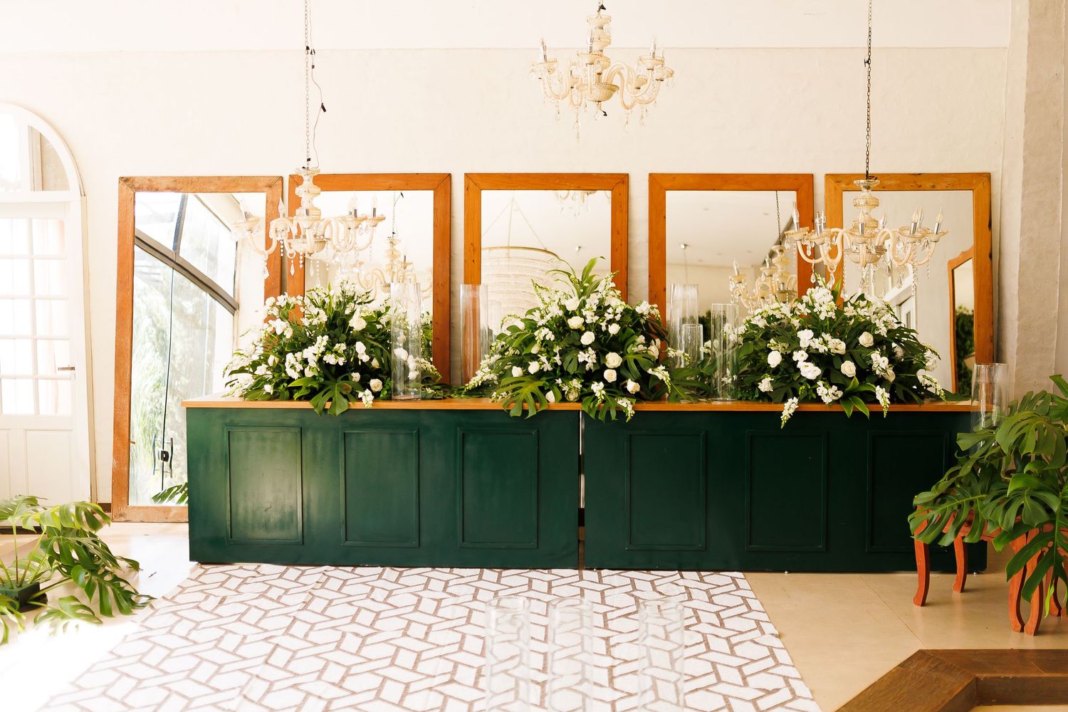 Elegant wedding venue with green bar, white floral arrangements, gold mirrors, and chandelier on patterned tile floor.