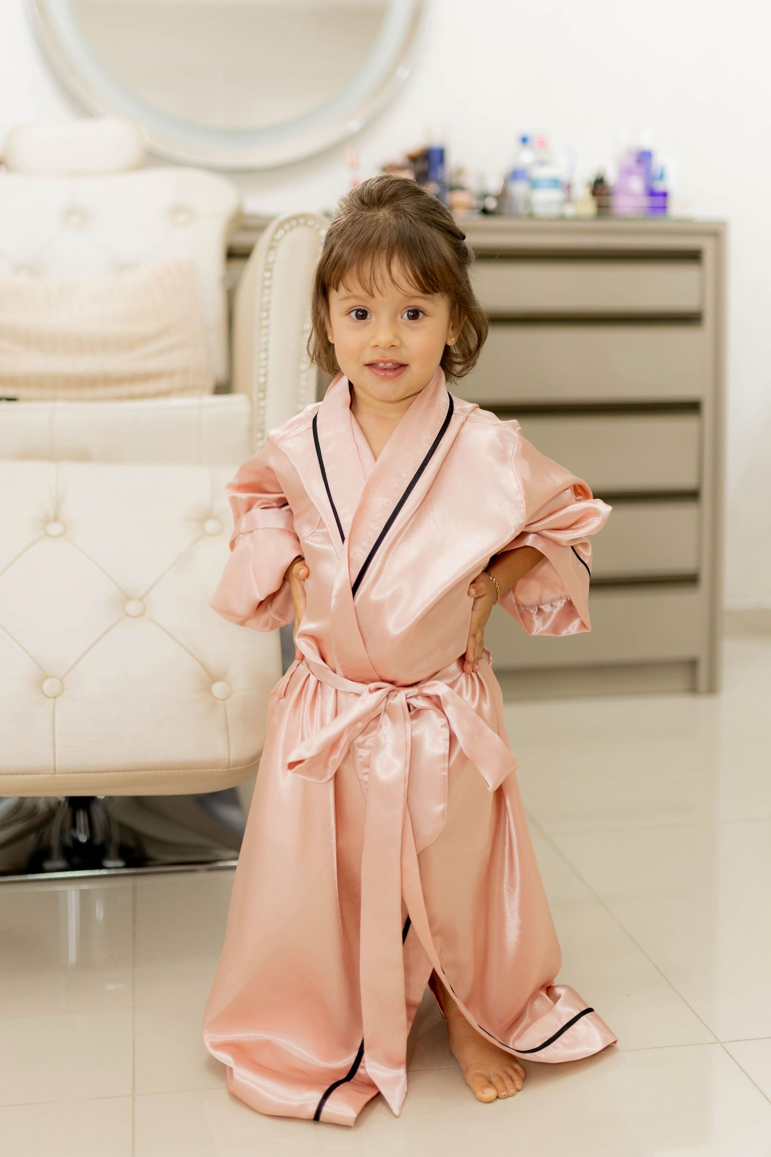 Adorable little girl wearing an oversized pink satin robe with navy trim, posing barefoot in a bright, elegant room.