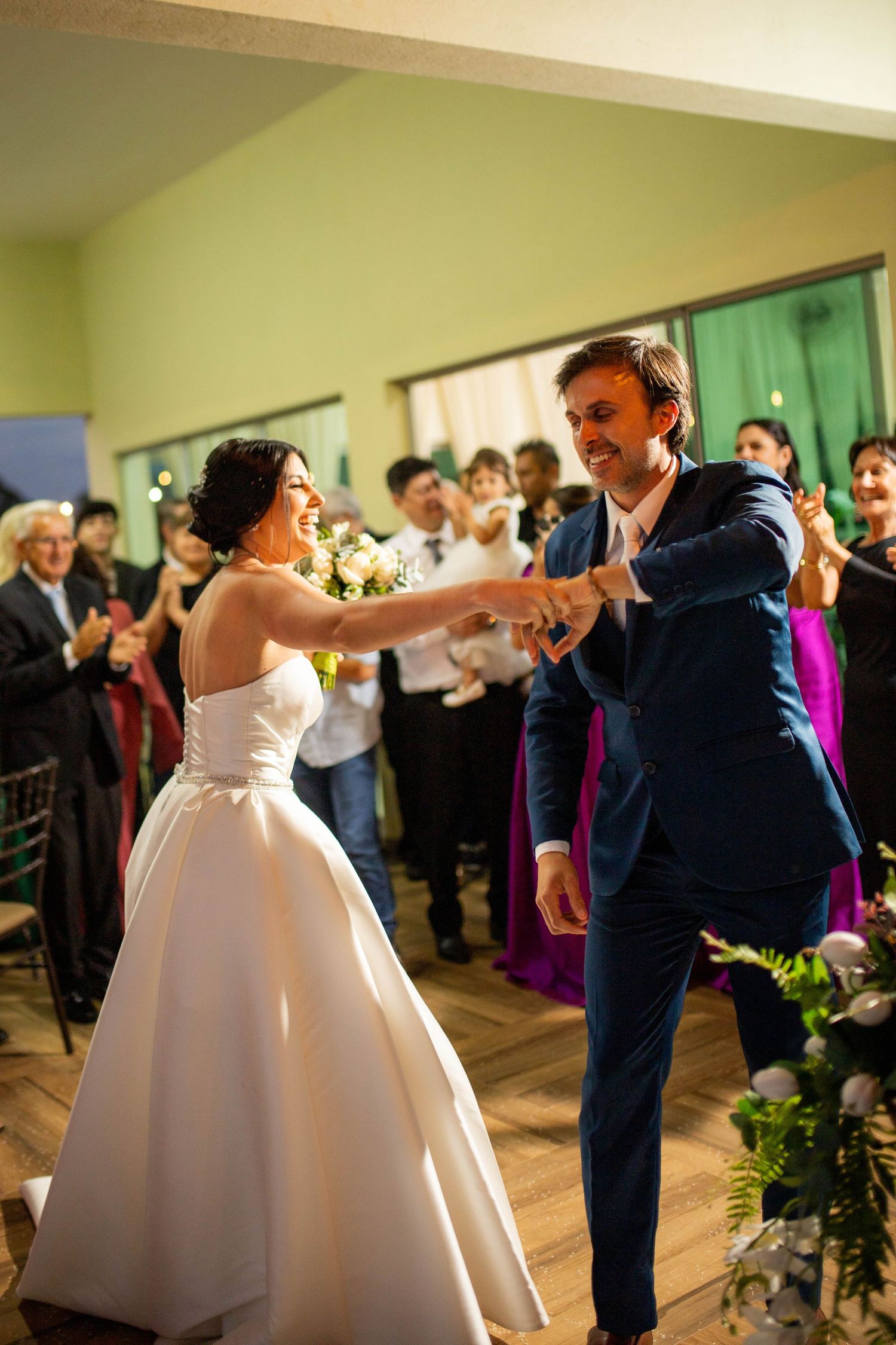 Bride and groom share their first dance at wedding reception, guests clapping in background.