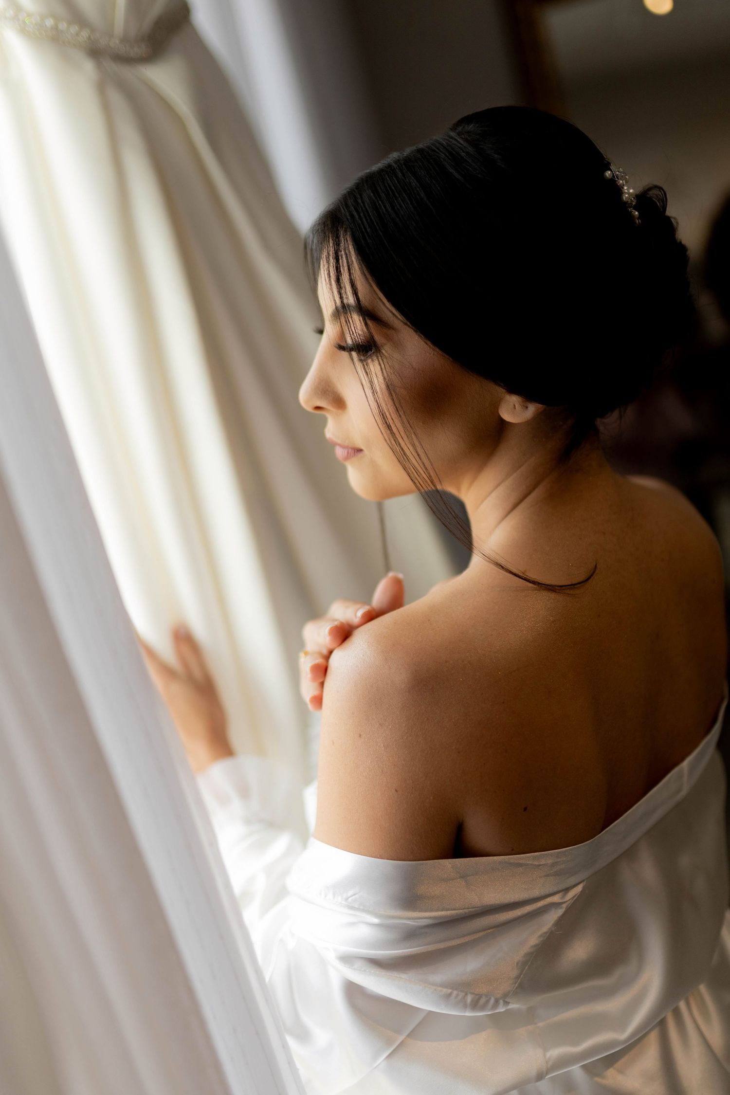 Bride in white satin robe gazes at wedding dress by window, showcasing elegant updo and bare shoulders.