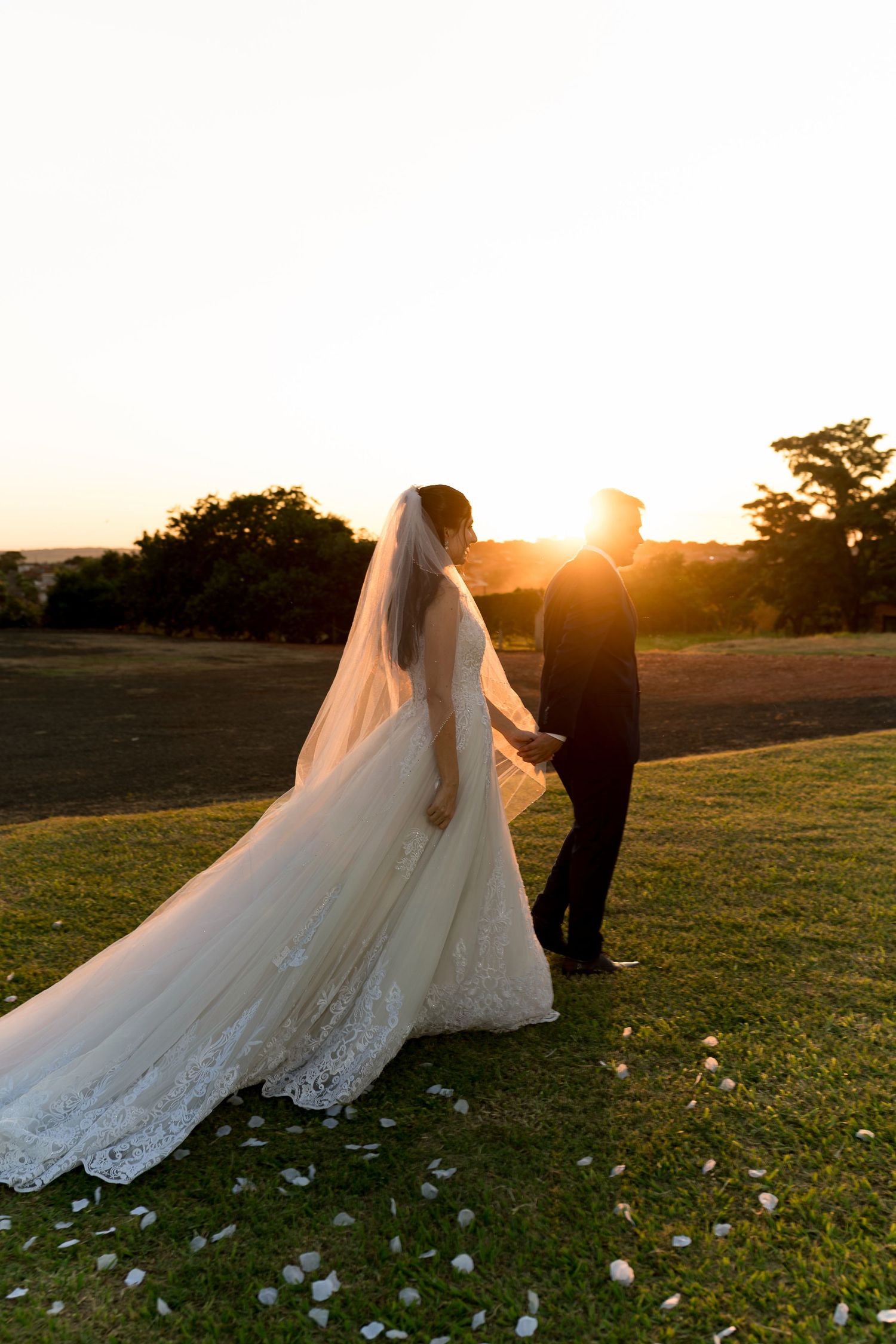Bride and groom holding hands at golden hour sunset on green lawn, long lace veil trailing behind.