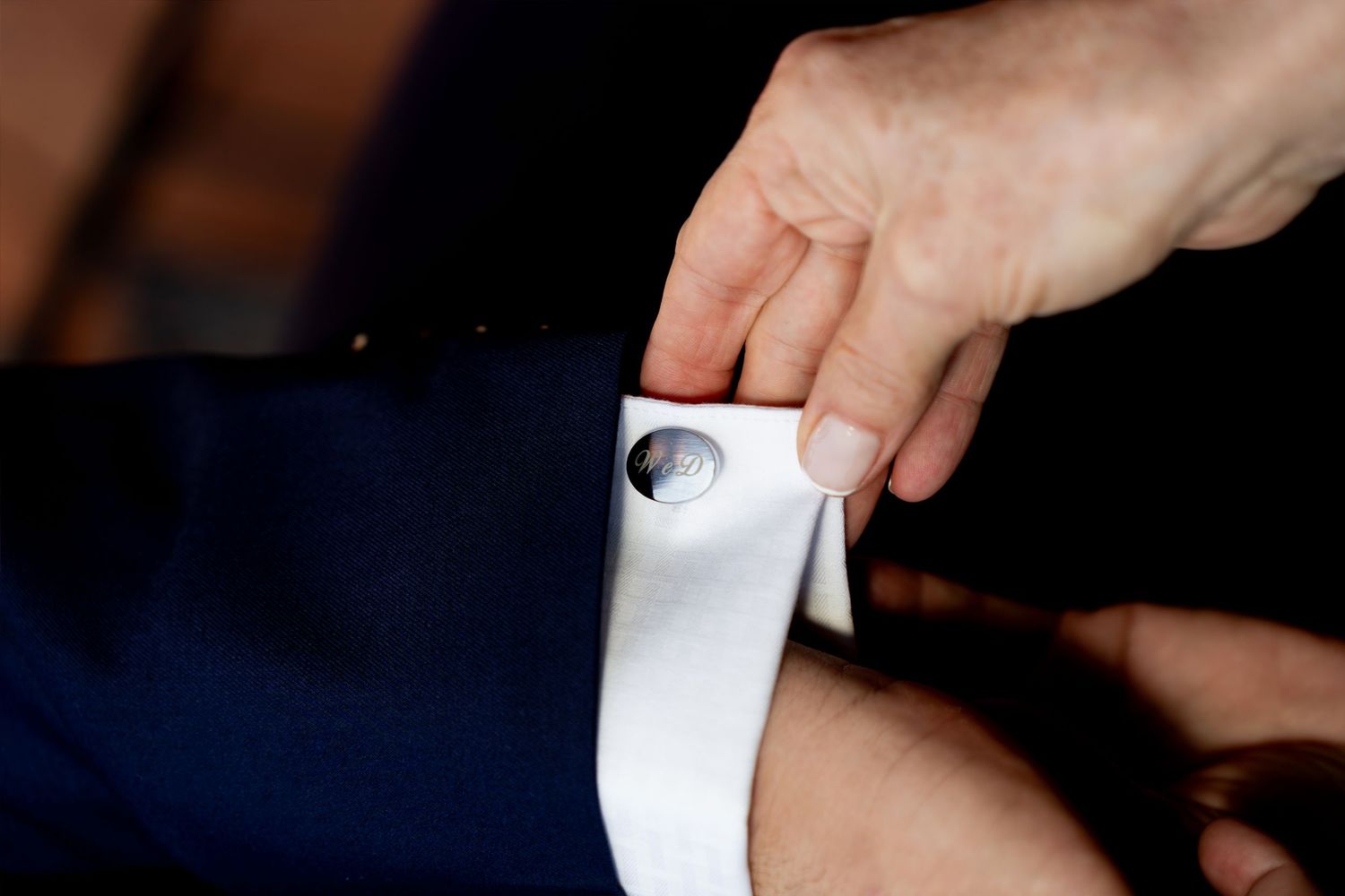 Close-up of hands fastening a silver cufflink on a white dress shirt sleeve with a navy suit jacket.