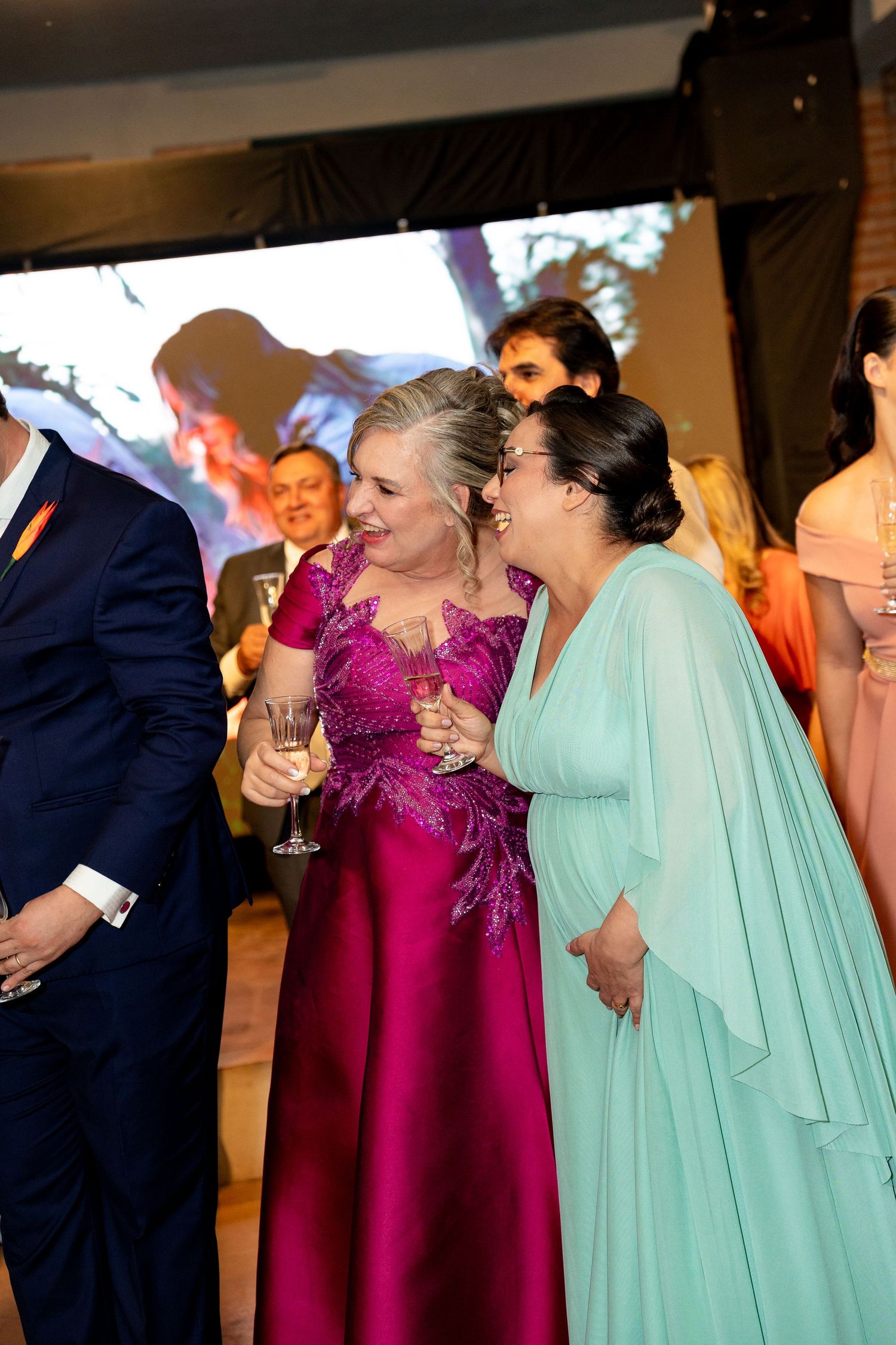 Two elegant women in formal gowns laughing together at a wedding reception, one in magenta and one in mint green.