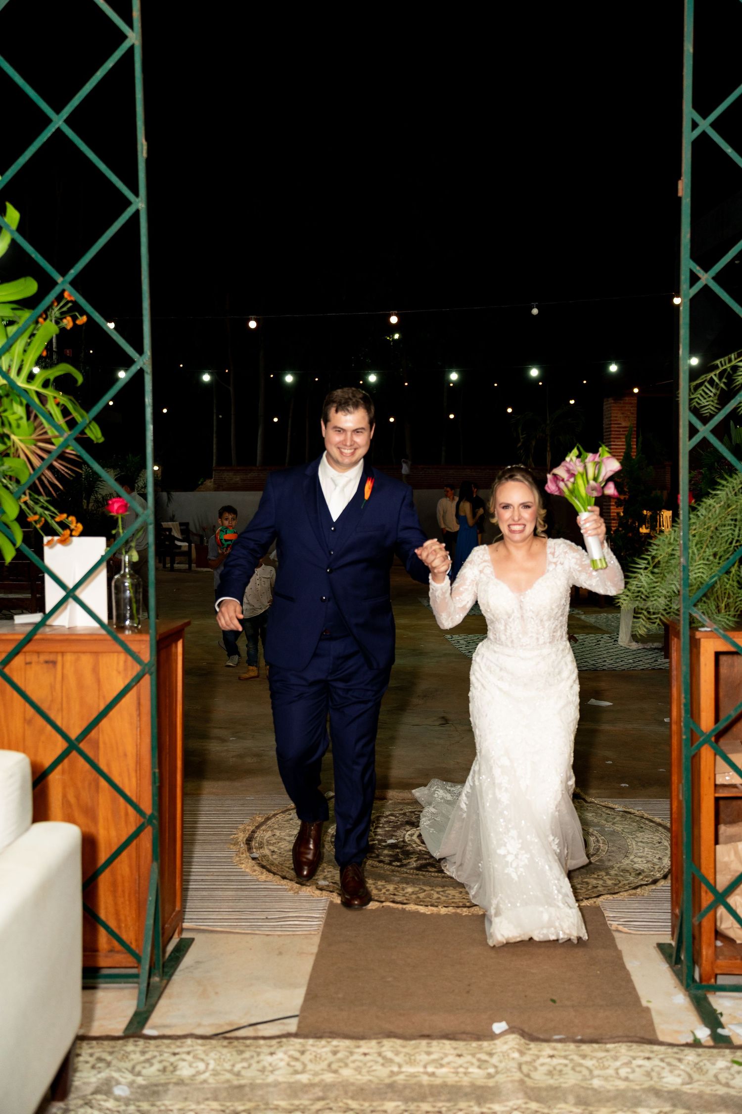 A joyful bride and groom walk hand-in-hand at their outdoor evening wedding reception, she holds a colorful bouquet aloft.