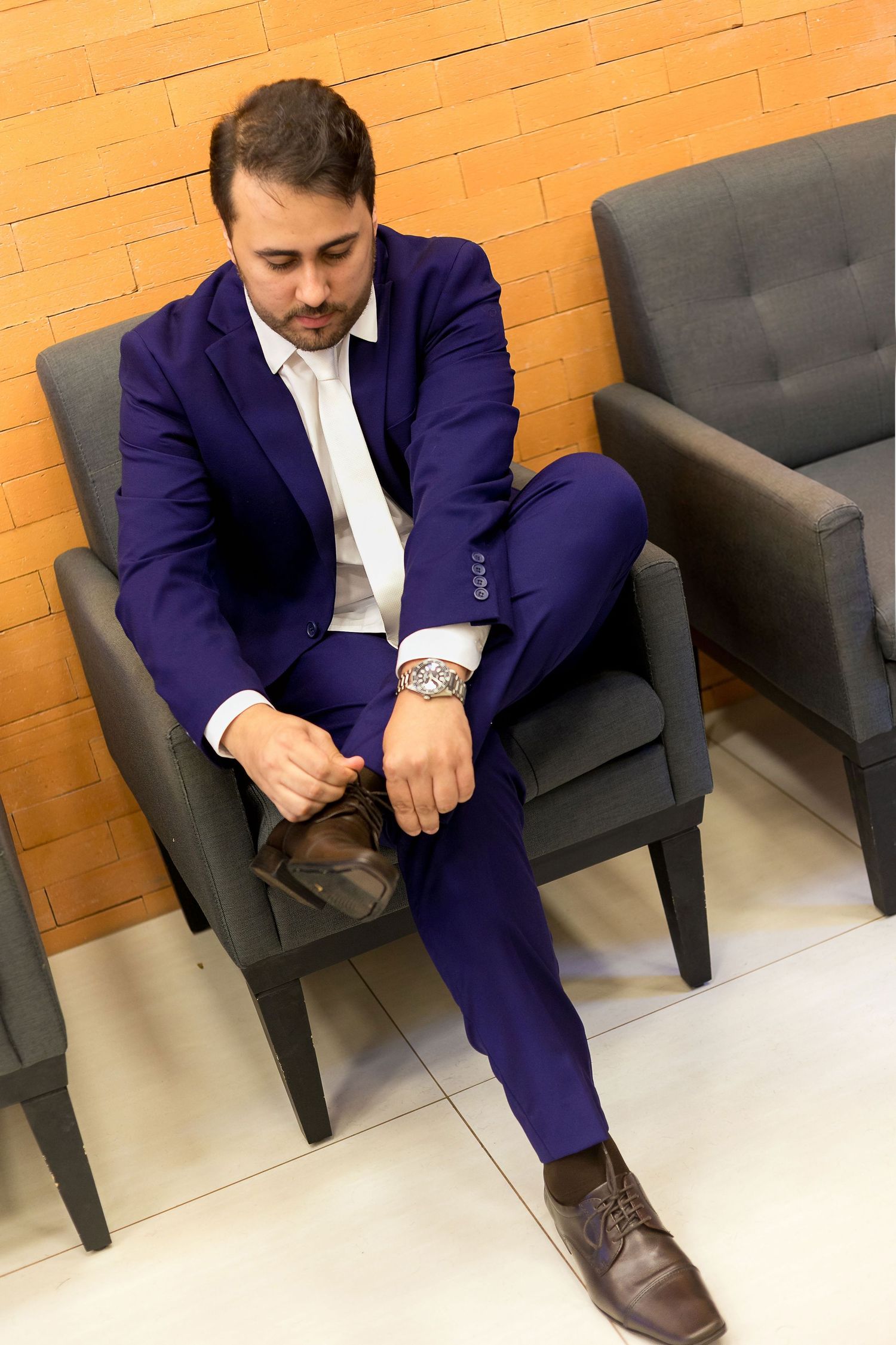 Man in blue suit adjusting brown dress shoes while seated in gray chair against brick wall.