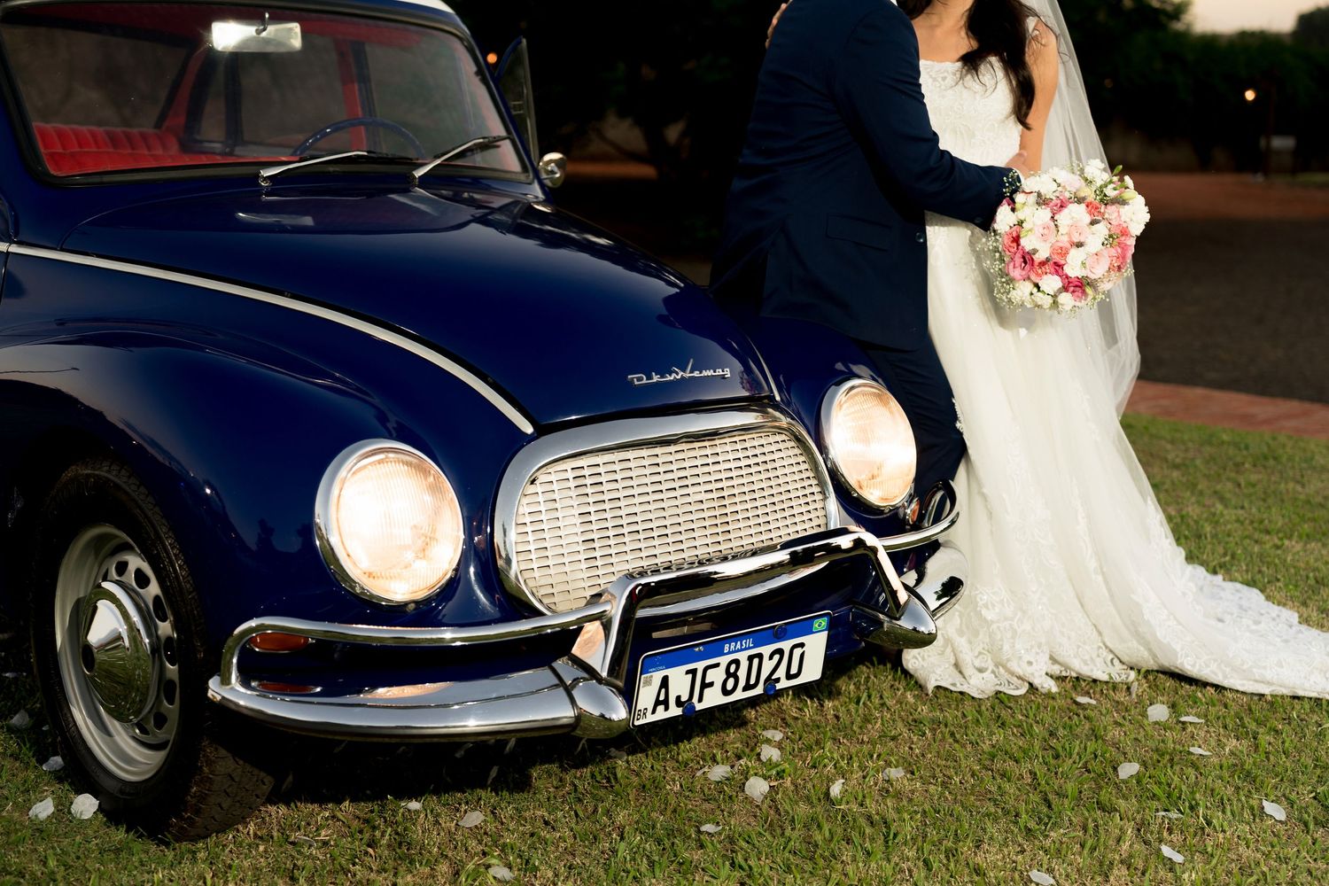 Newlywed couple posing beside a classic vintage navy blue car with a bouquet at an outdoor wedding.