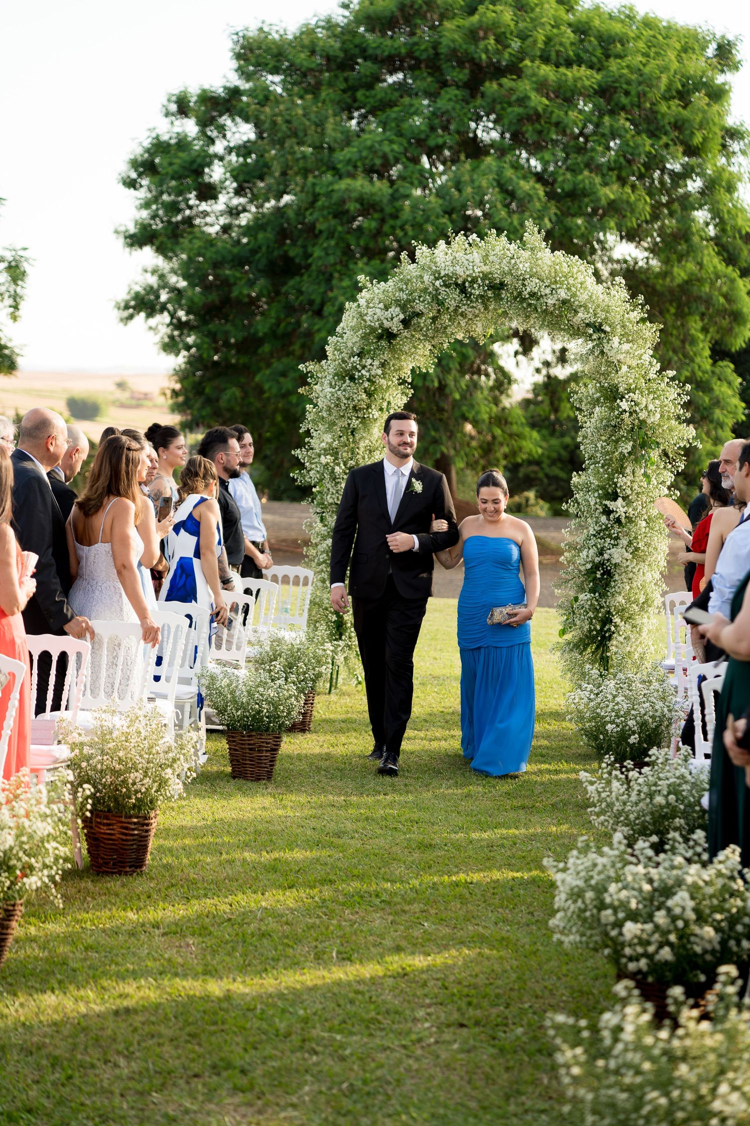Outdoor wedding ceremony with guests seated in white chairs, floral arch, and couple walking down the aisle on green lawn.