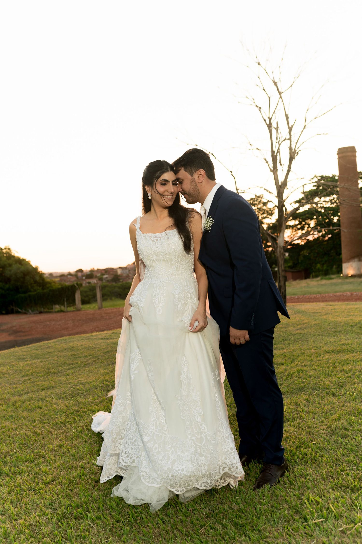 Bride in white lace gown and groom in navy suit share a romantic moment on green lawn at sunset.